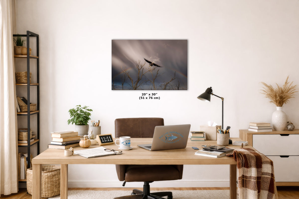 A wall art print depicting a bird perched on bare tree branches against a dramatic cloudy sky with a faint rainbow, displayed in a styled home office setting above a wooden desk.