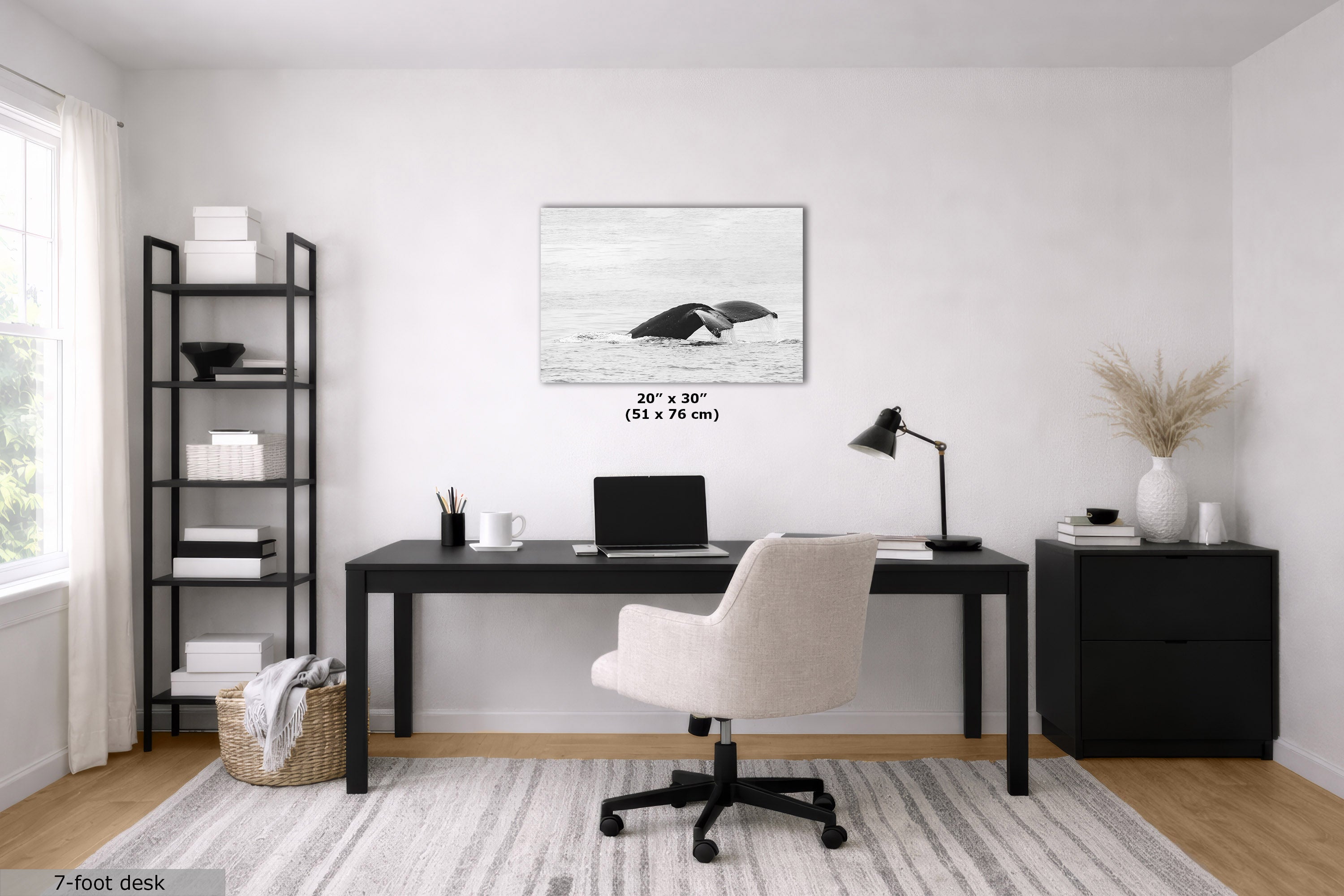 A modern home office setup featuring a black 7-foot desk, a cream swivel chair, a black shelving unit, and a black side cabinet. The focal point on the white wall is a black and white framed print of a whale, with dimensions '20" x 30" (51 x 76 cm)' printed below it.