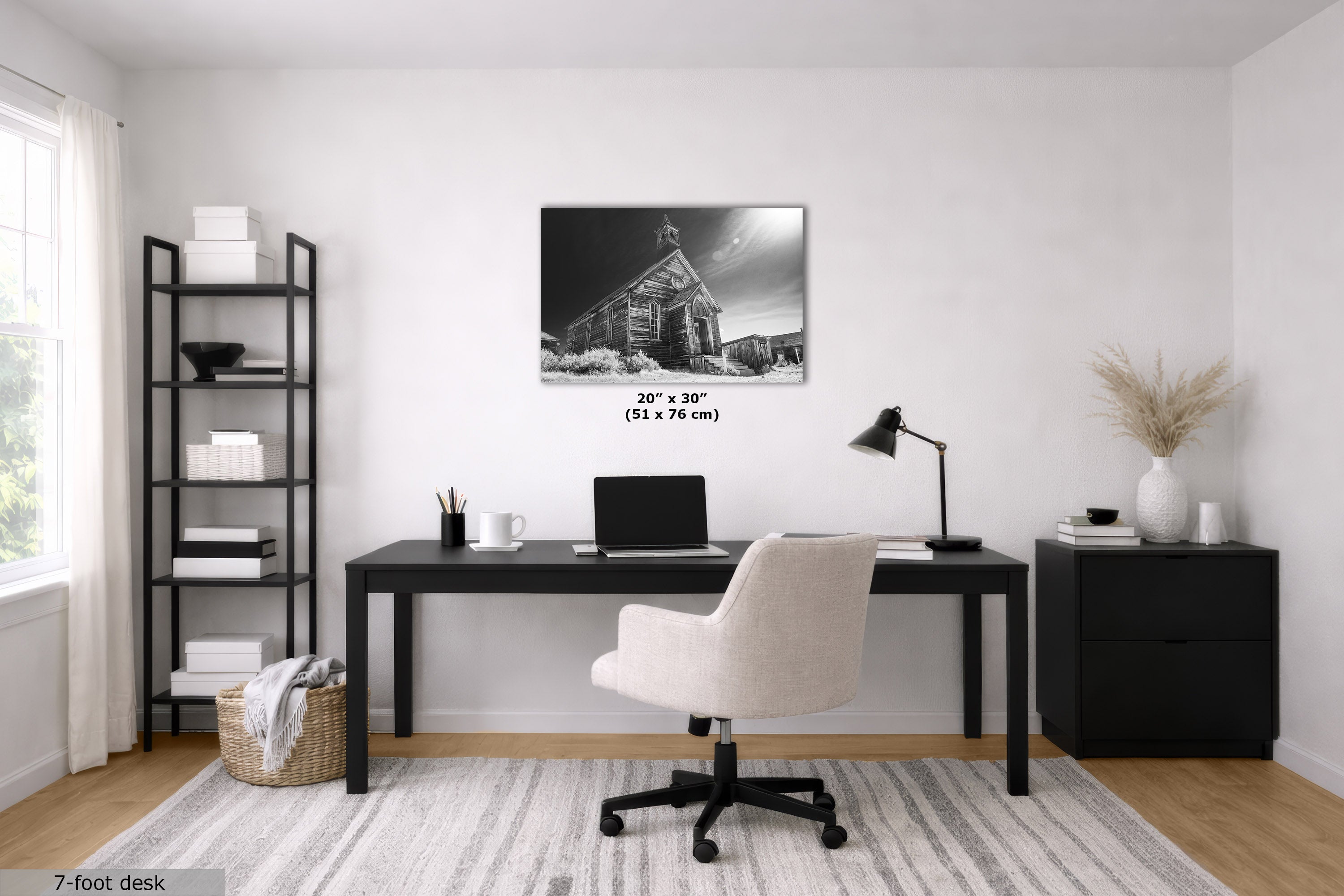 A styled home office scene featuring a black-framed black and white art print, labeled '20" x 30" (51 x 76 cm)', hung on a white wall above a dark desk and a cream office chair.