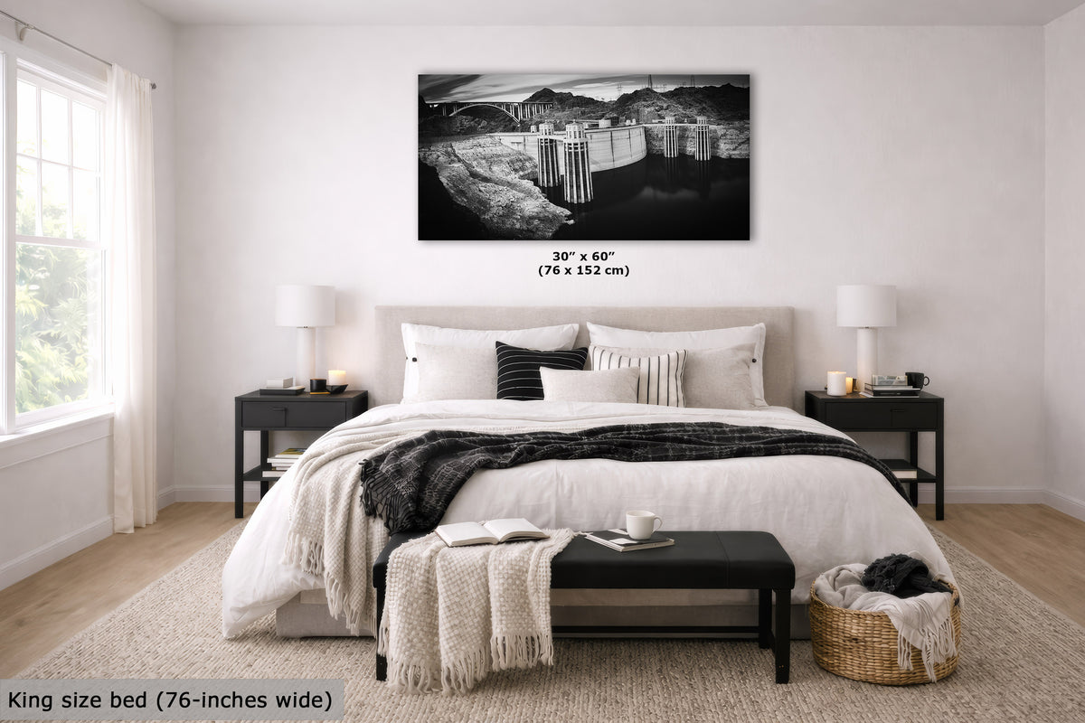 A black and white landscape wall art print displayed above a neatly made bed in a modern bedroom. The print shows a dam and reservoir scene.
