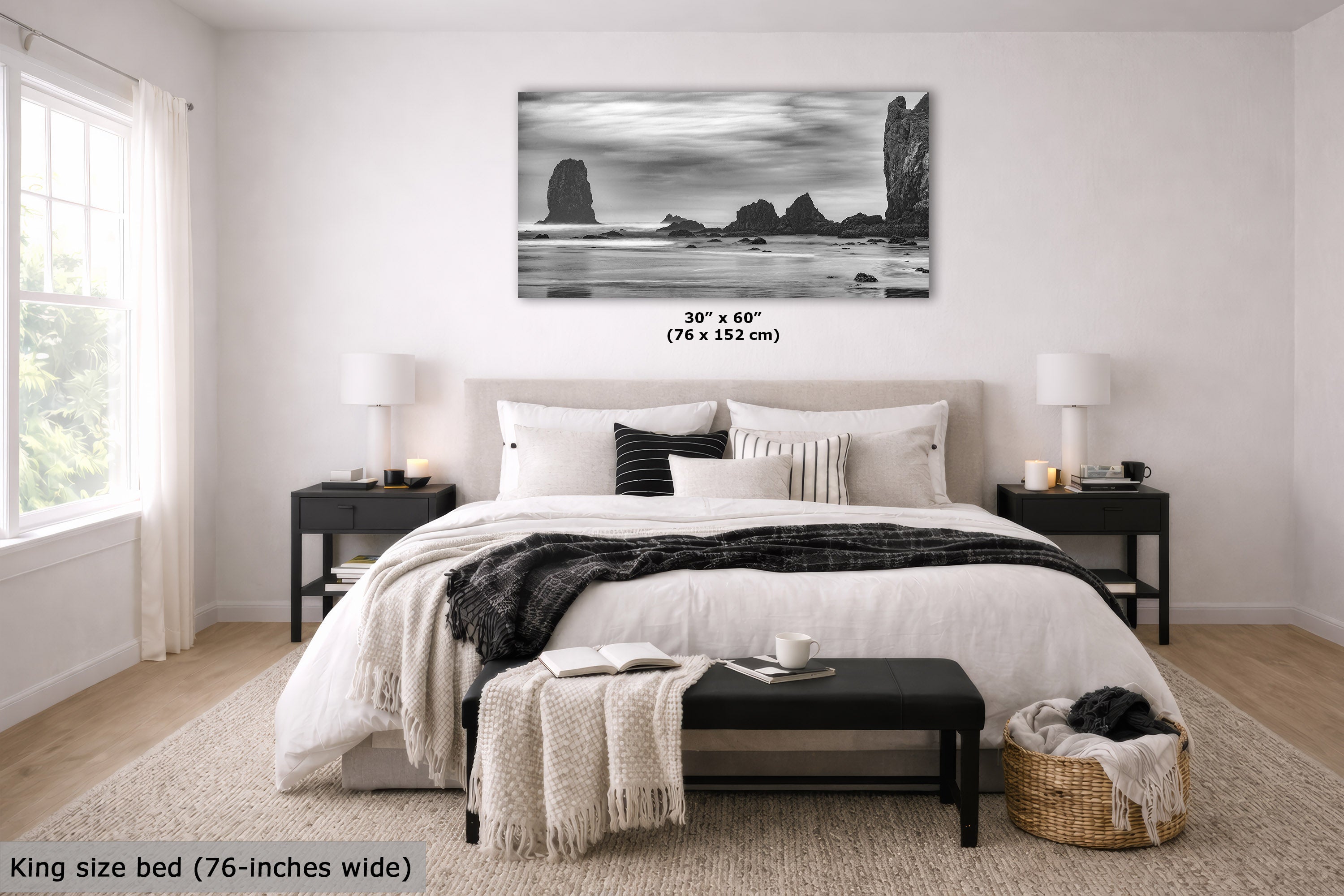 A black and white coastal rock landscape wall art print is displayed above a neatly made bed in a bright, modern bedroom. The print shows tall sea stacks and cliffs over the ocean.