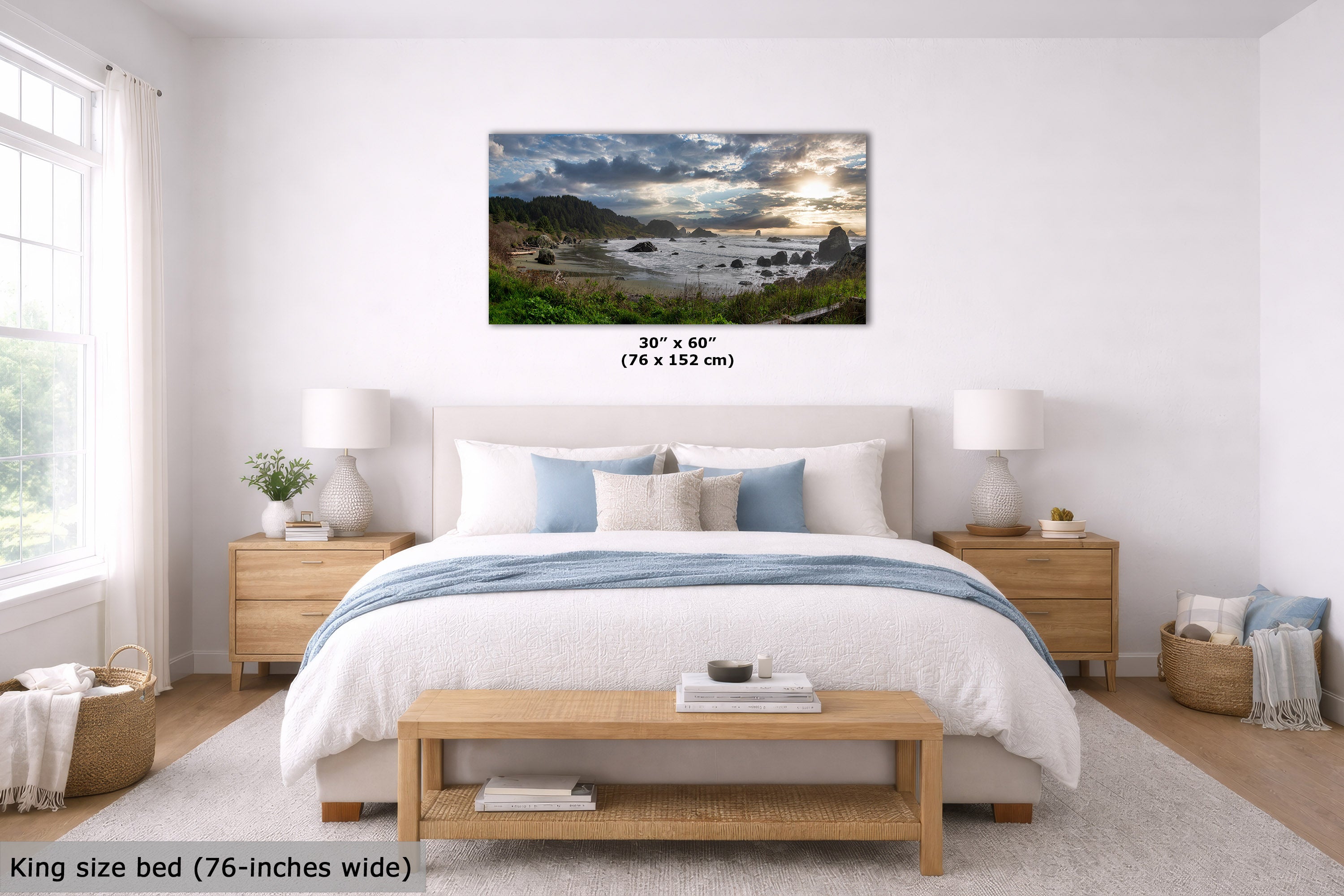 A framed coastal landscape wall art print hangs above a neatly made bed in a bright, modern bedroom. The print shows a rocky shoreline with dramatic clouds and warm sunlight.
