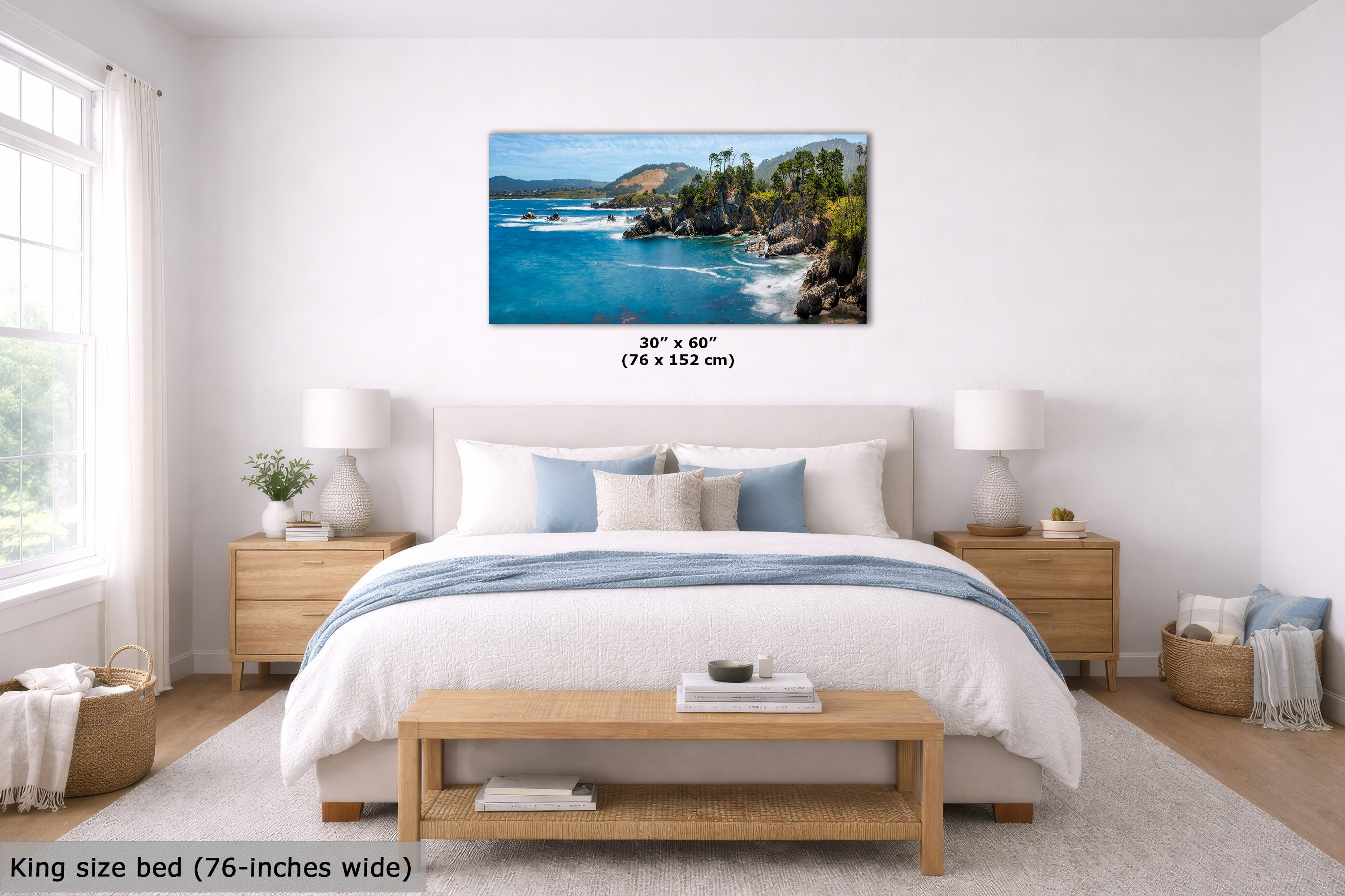 A framed coastal landscape wall art print hangs above a neatly made bed in a bright, modern bedroom. The artwork shows turquoise ocean water, rocky shoreline, green trees, and blue sky.