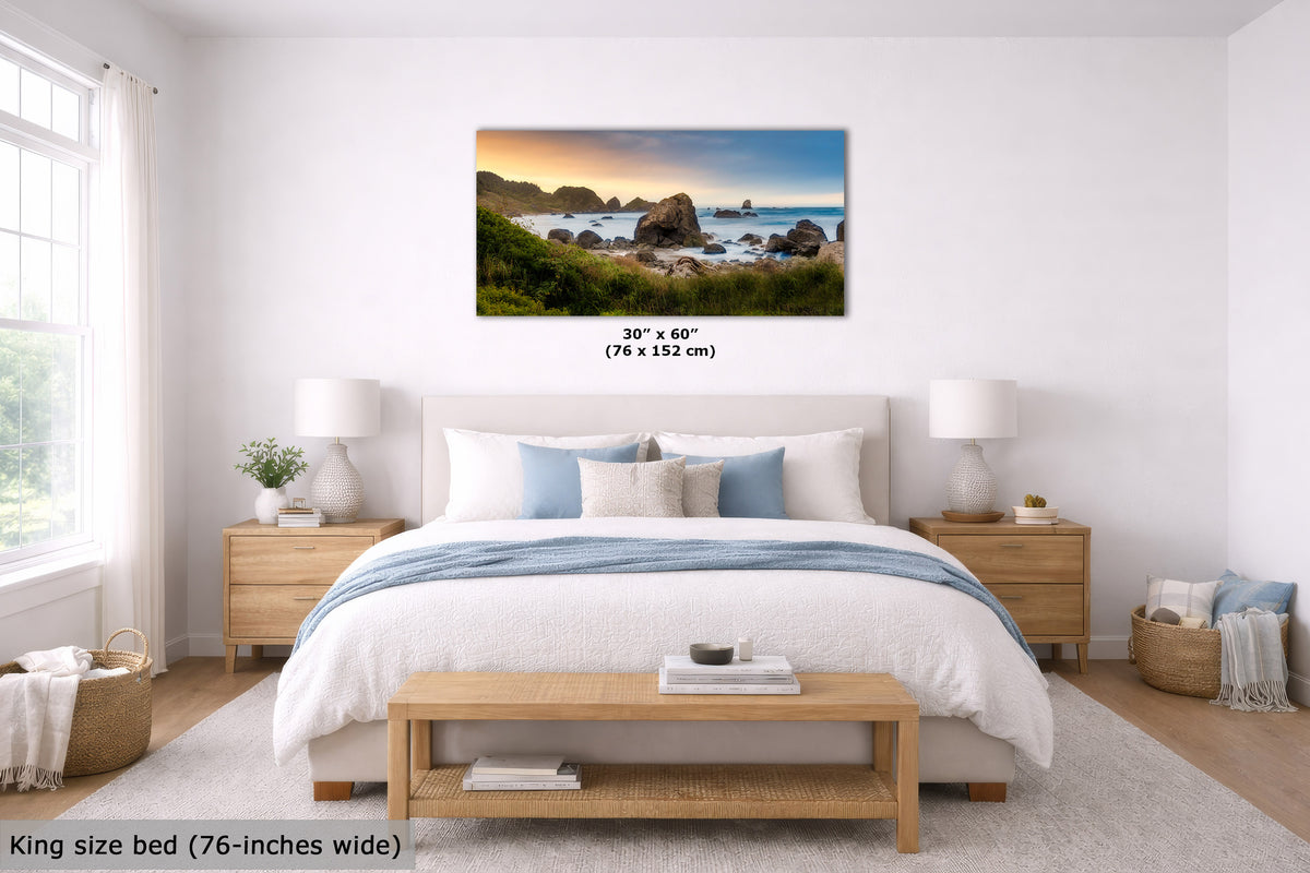 A coastal landscape wall art print hangs above a neatly made bed in a bright, modern bedroom. The print shows a rocky beach at sunset with blue water and green cliffs.
