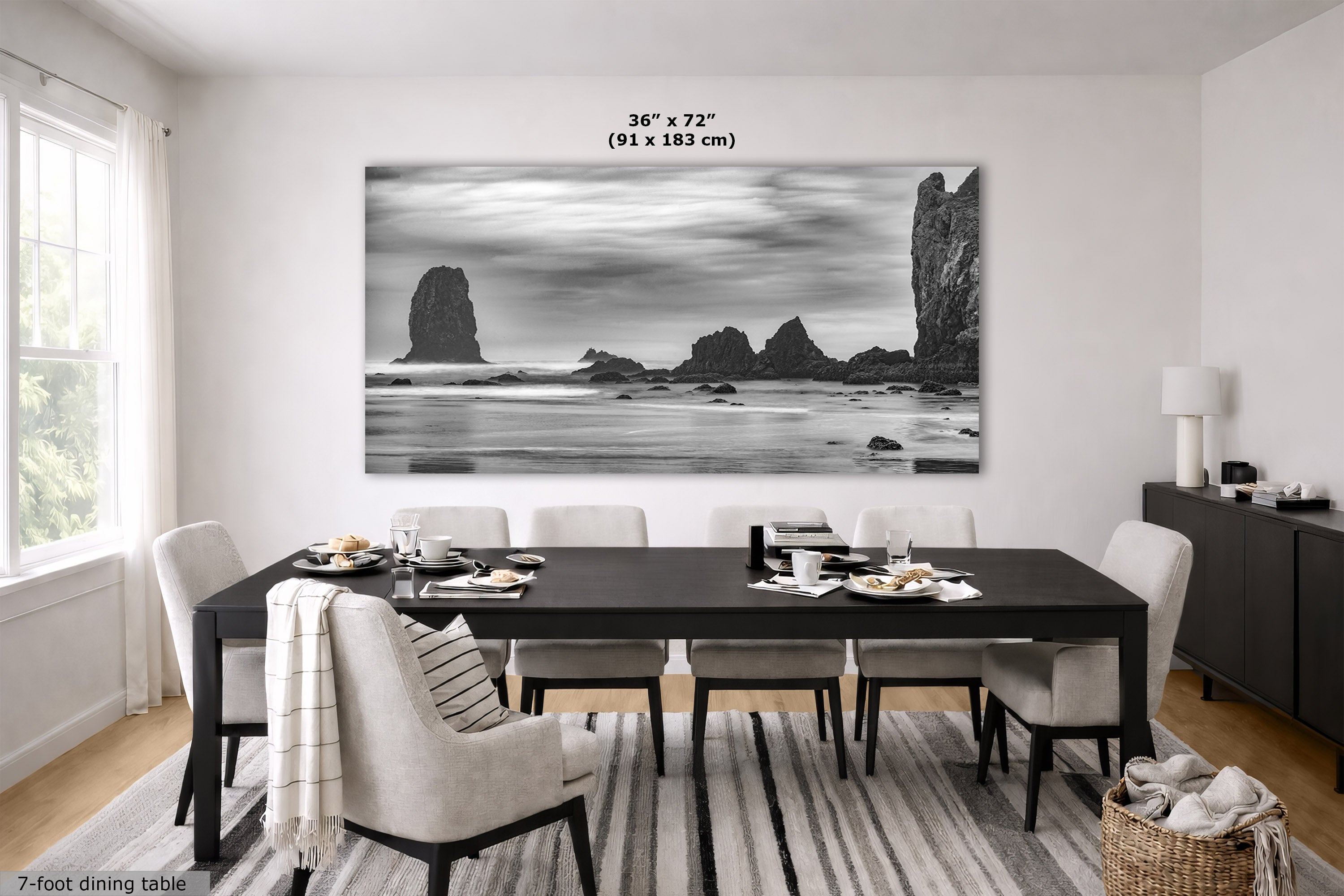 A large black and white coastal rock formation wall art print hangs above a dark wood dining table with upholstered chairs in a bright, modern dining room. The print includes size text reading 36 x 72 inches (91 x 183 cm) and a small label at bottom left reading 7-foot dining table.