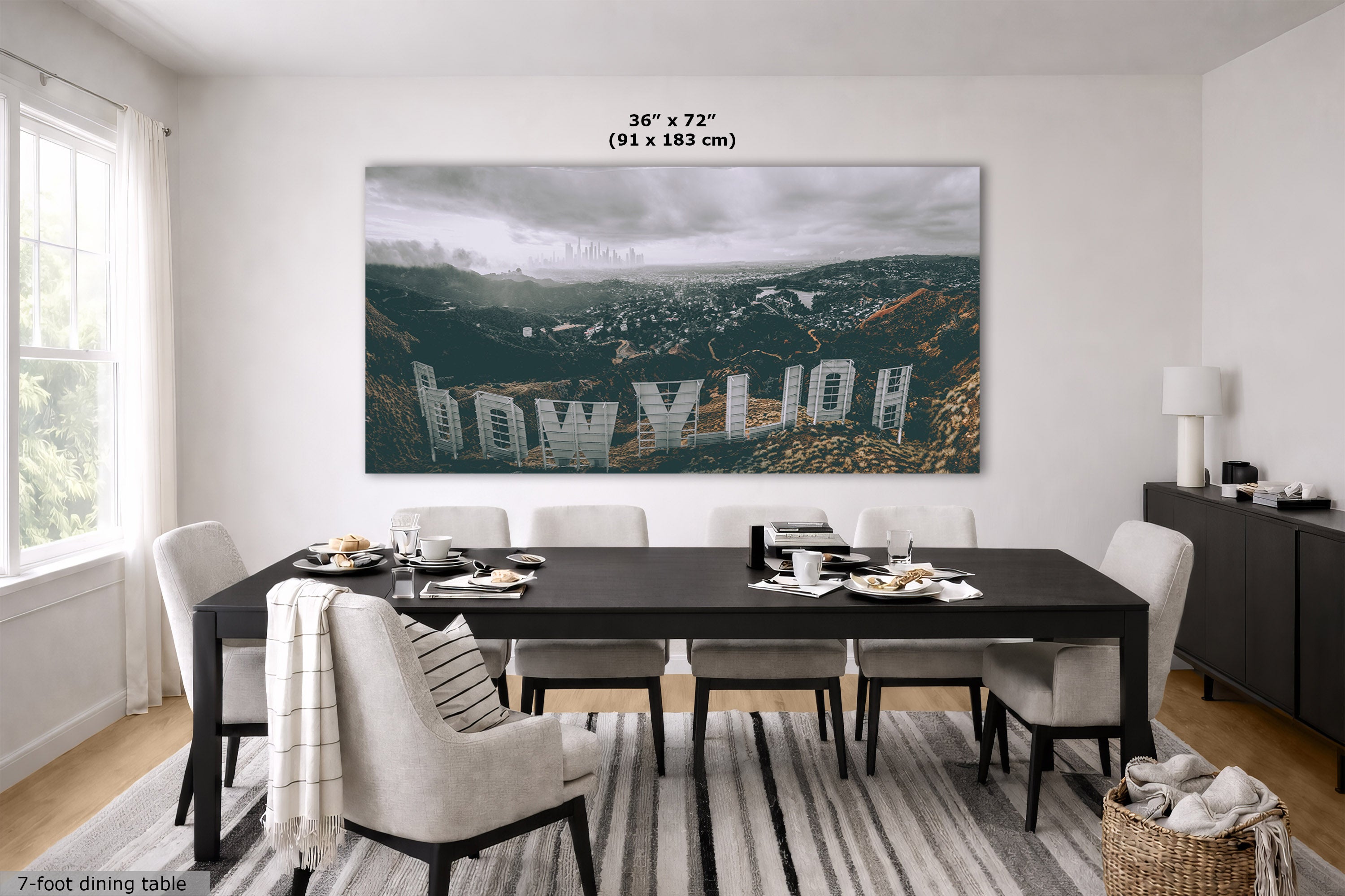 A large rectangular wall art print showing a moody city skyline over hills with the word 'HOLLYWOOD' across it, displayed above a dining table in a bright modern room. The print includes size text reading 36 x 72 in (91 x 183 cm).