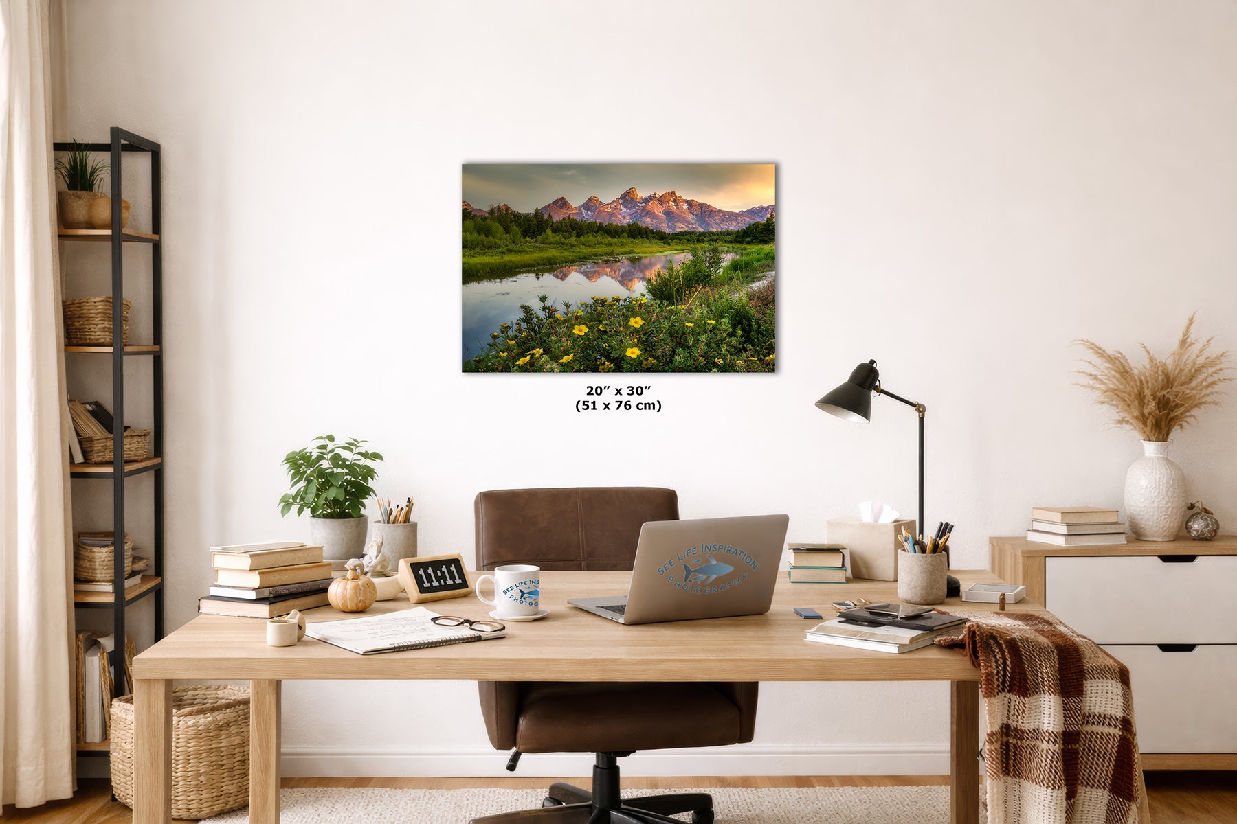 A framed wall art print showing the Grand Teton mountain landscape reflected in a calm river with wildflowers, displayed above a wooden desk in a styled home office. The print includes size text reading 20 x 30 inches (51 x 76 cm).