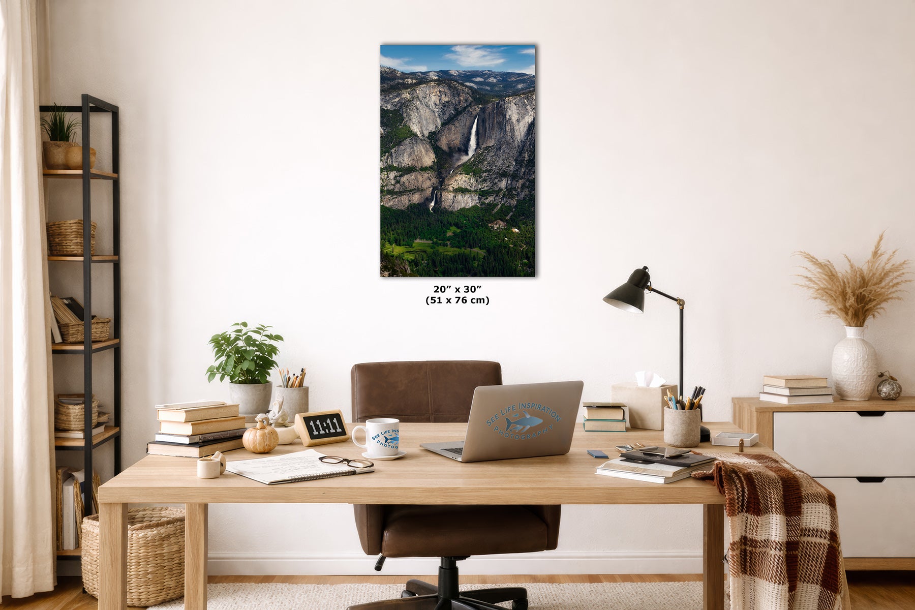 A vertical wall art print depicting a scenic waterfall and rocky mountain landscape, styled in a modern home office setting above a wooden desk with a laptop, lamp, and chair.