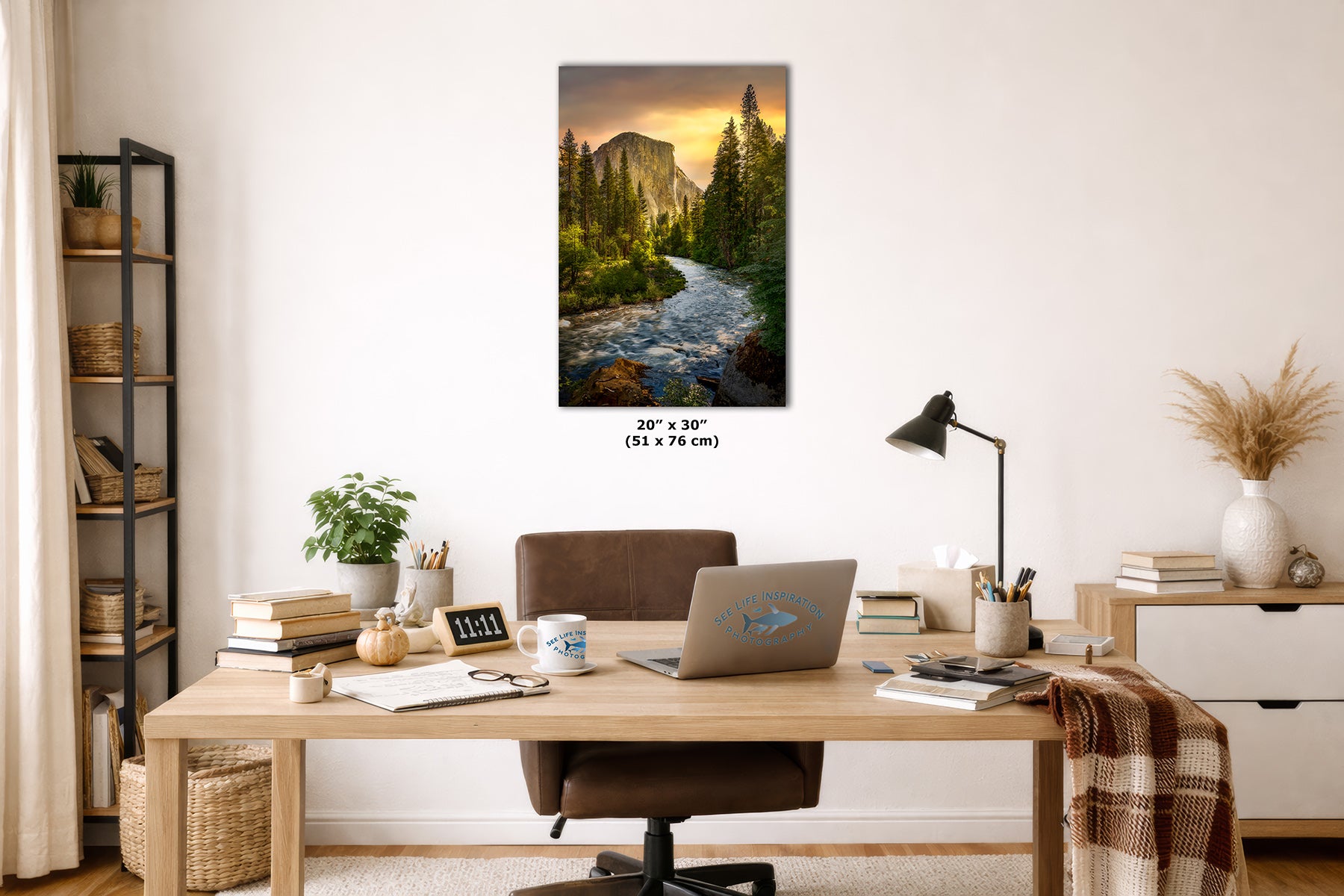 A vertical landscape art print depicting a rushing Merced River through the Yosemite forest toward El Capitan mountain at sunset, displayed on a white wall above a wooden desk in a styled home office setting.