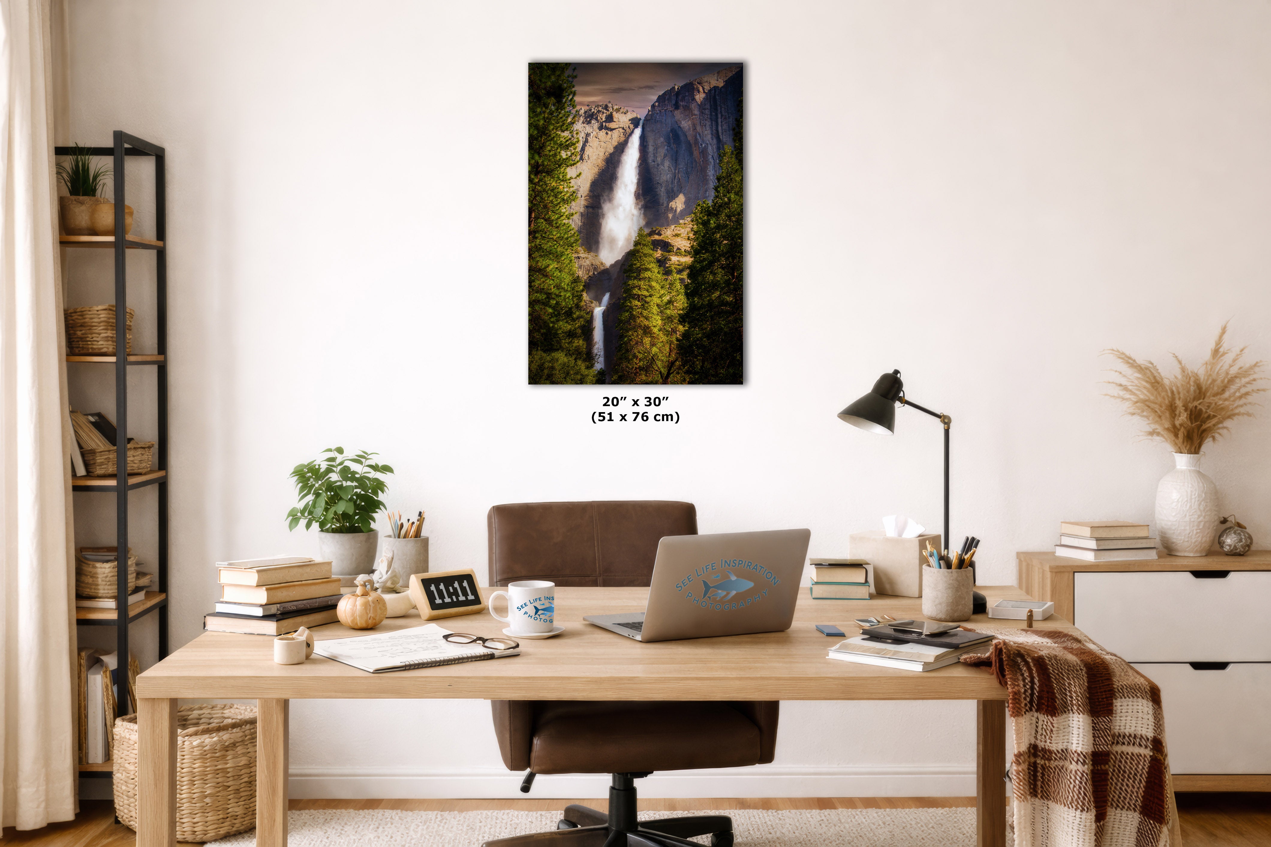 A vertical wall art print depicting a waterfall in a forest, displayed in a styled home office setting above a wooden desk with a laptop, lamp, and other office accessories. The print includes size text below it reading 20x 30 inches (51 x 76 cm).