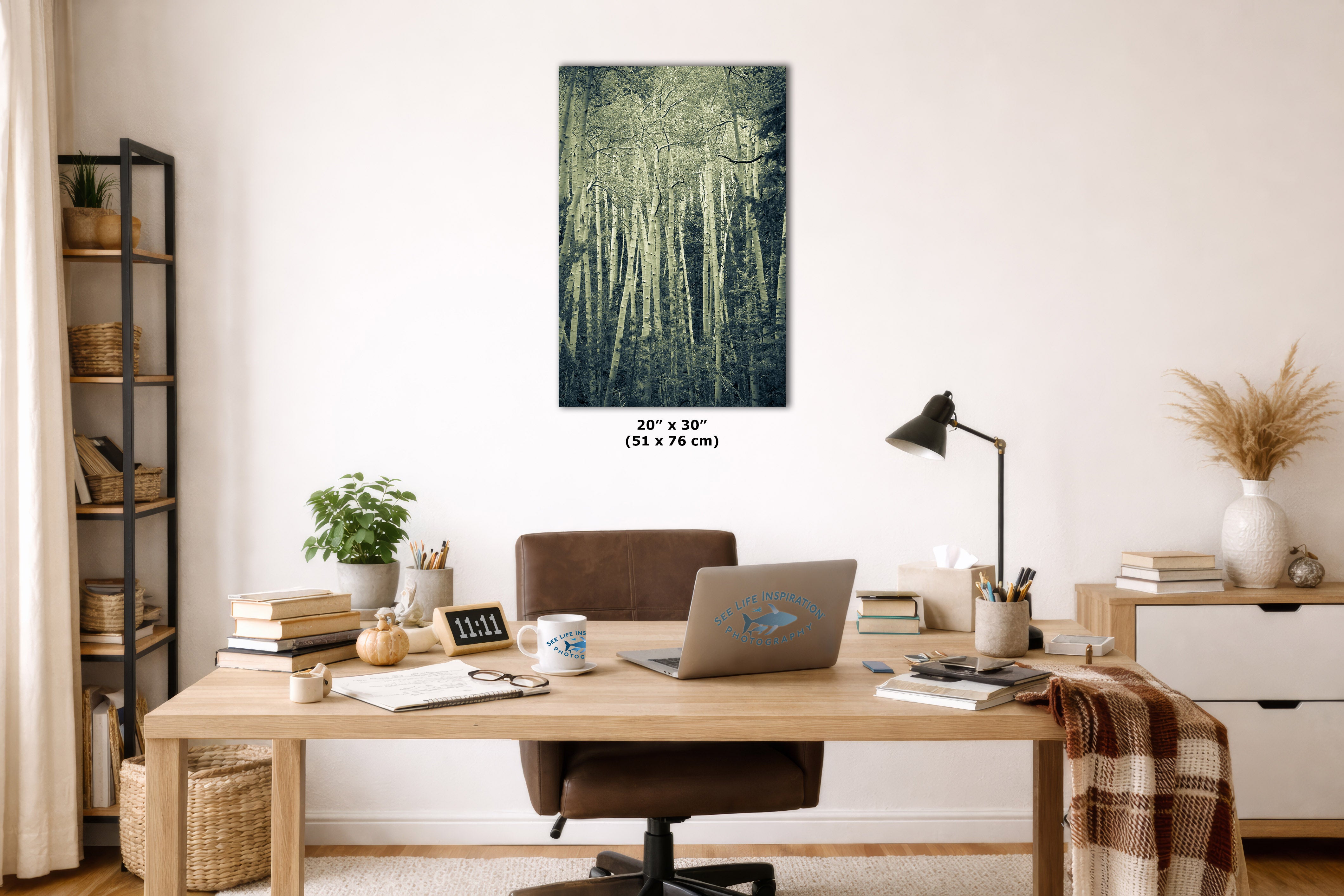 A vertical canvas wall art print depicting a dense forest with cool green and blue tones, styled in a modern home office setting above a wooden desk with a laptop, lamp, and books.