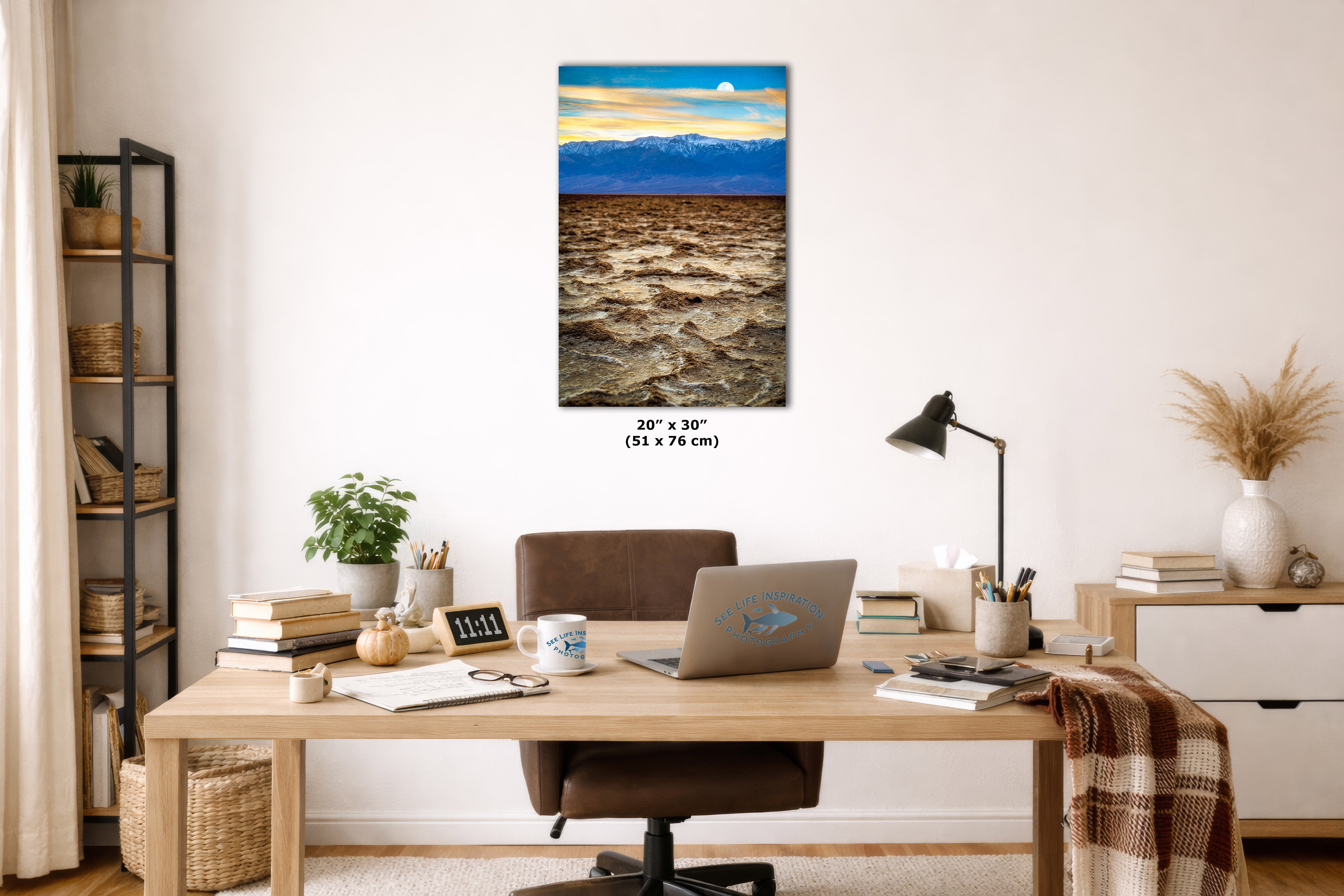 A vertical canvas wall art print depicting a desert landscape under a blue sky, styled in a modern home office setting above a wooden desk with a laptop and chair.