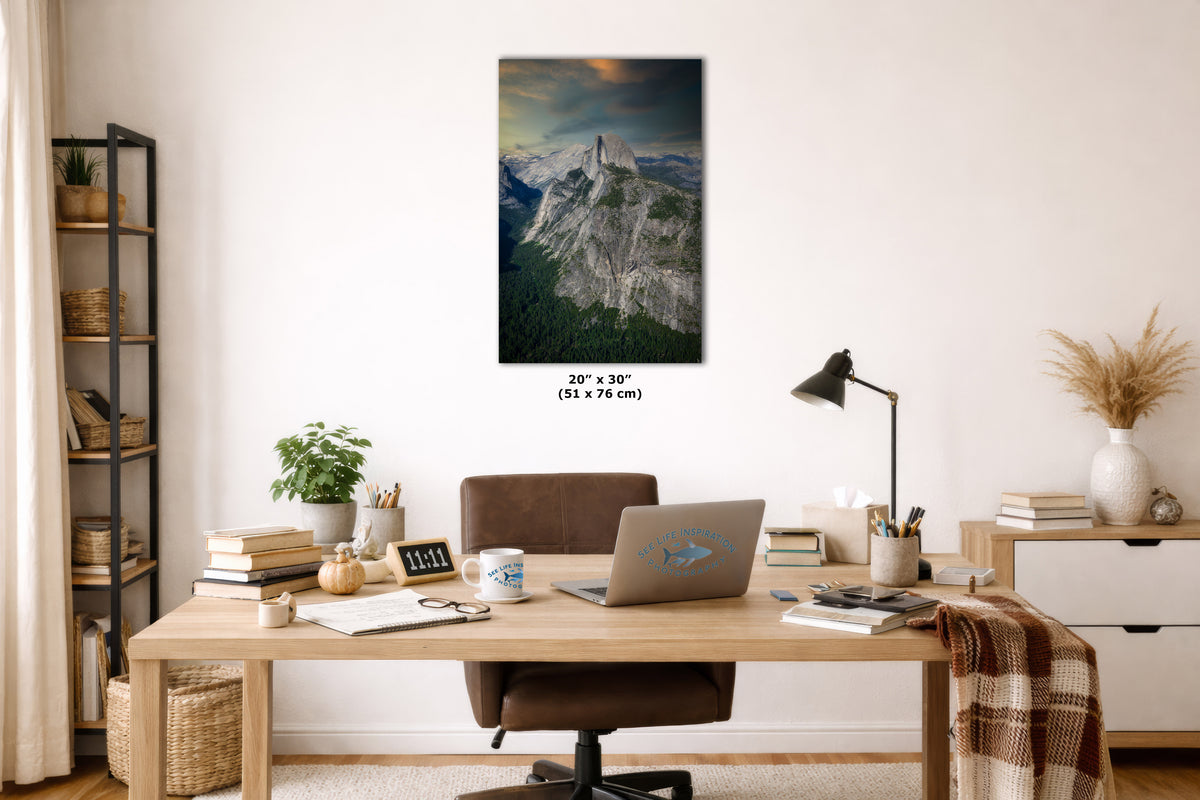 A vertical canvas wall art print depicting a dramatic mountain landscape with a glowing sunset, displayed on a white wall above a wooden desk in a modern home office setting.