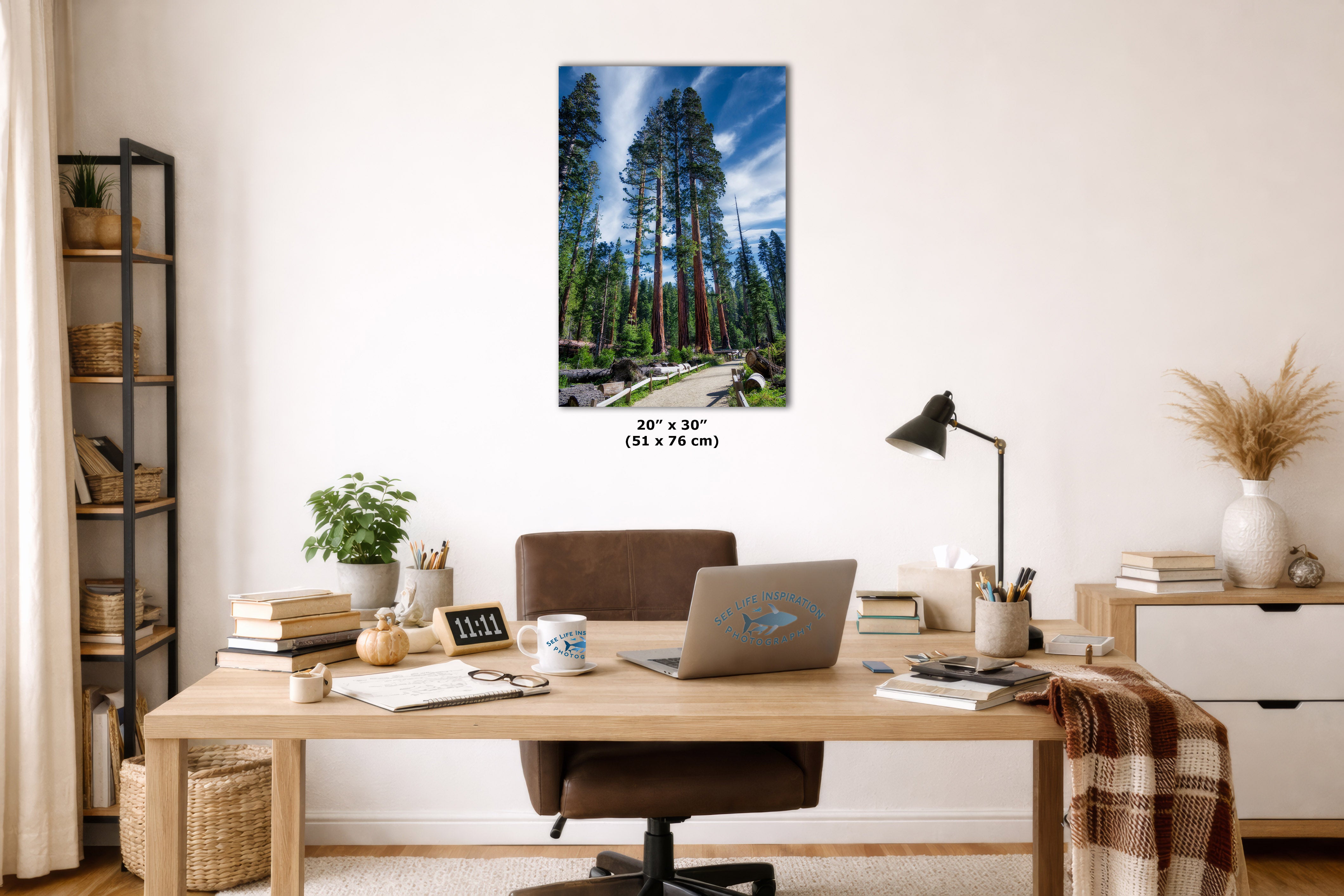 A vertical canvas print of a forest scene with tall trees and a blue sky, displayed on a wall above a wooden desk in a modern home office setting.