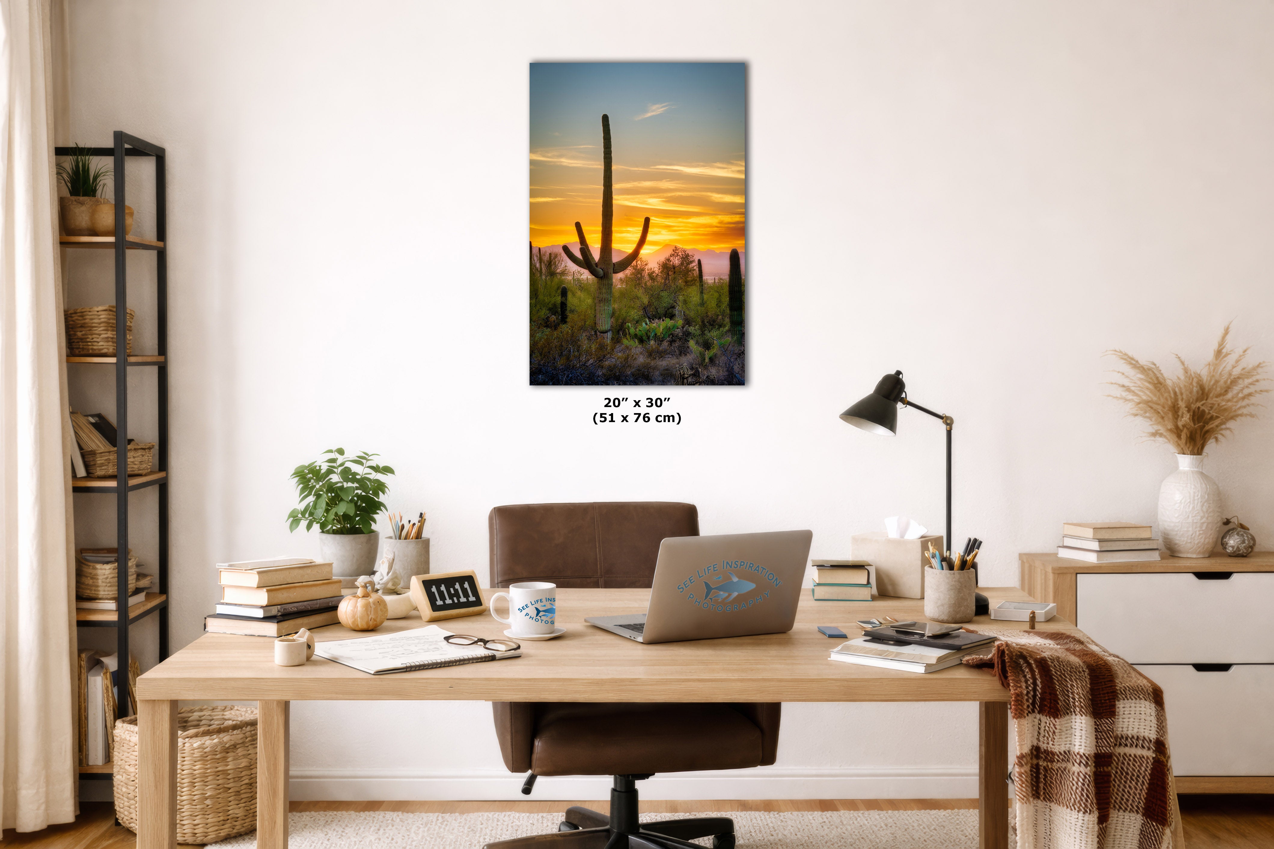 A vertical canvas wall art print depicting a desert sunset with a saguaro cactus, displayed on a white wall above a wooden desk in a styled home office setting.