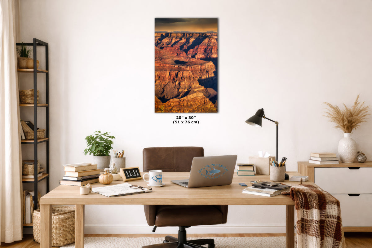 A vertical canvas wall art print depicting the Grand Canyon at sunset, styled in a modern home office setting above a wooden desk with a laptop, lamp, and books.