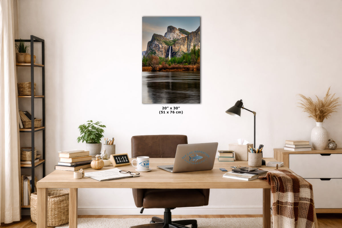 A vertical wall art print depicting a waterfall in a forest with a river in the foreground, displayed in a styled home office setting above a wooden desk with a laptop, lamp, and other office accessories. The print includes size text below it reading 20x 30 inches (51 x 76 cm).