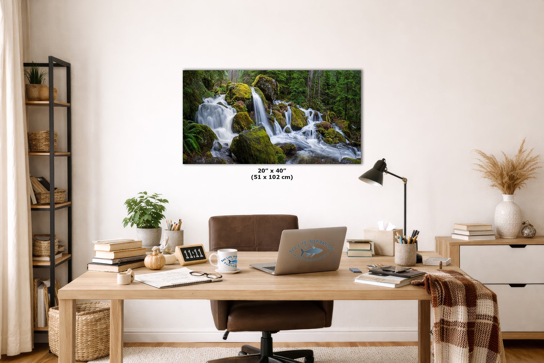 A framed wall art print showing a lush green forest waterfall, displayed in a styled home office setting above a wooden desk with a laptop, lamp, and other office accessories. The dimensions '20" x 40" (51 x 102 cm)' are printed below the artwork.