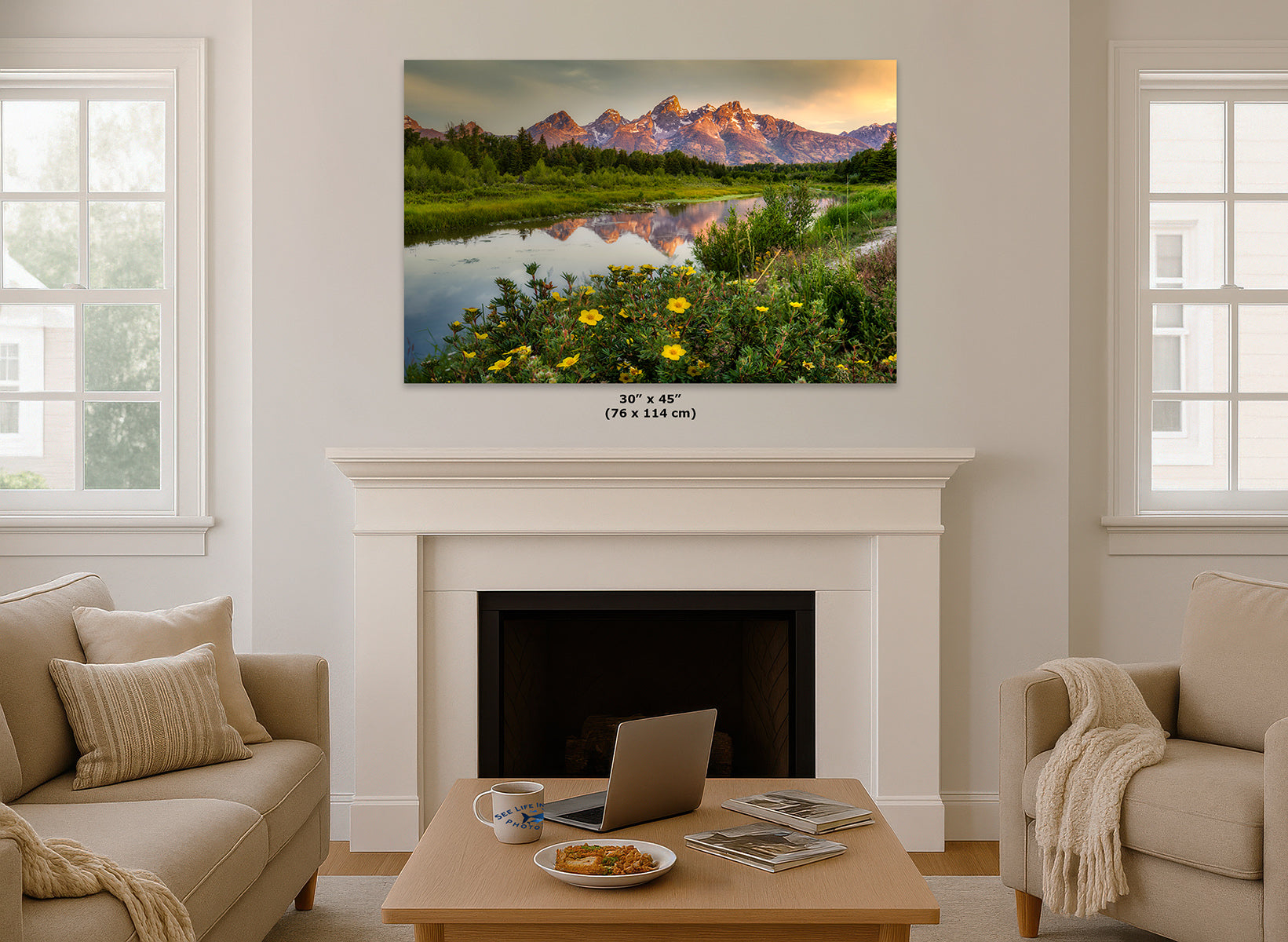 A wall art print depicting the Grand Teton Mountains with a reflective lake and yellow wildflowers, displayed above a white fireplace in a bright living room. Small text below the print reads 36 x 45 in (76 x 114 cm).