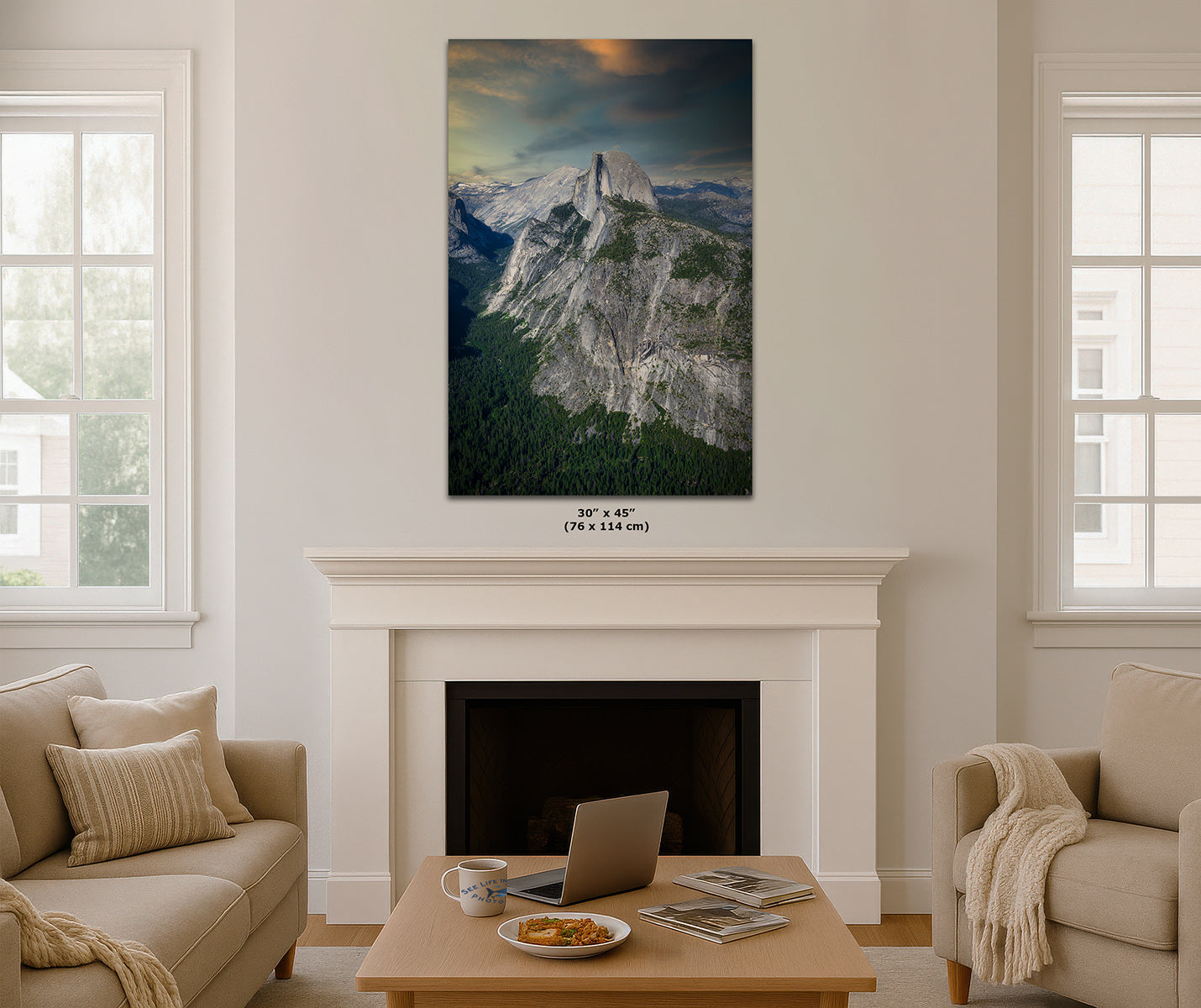 A vertical wall art print of Half Dome in Yosemite depicting a dramatic mountain landscape with a tall rocky peak, a blue sky, and a dark forest below, displayed above a white fireplace in a modern living room setting.