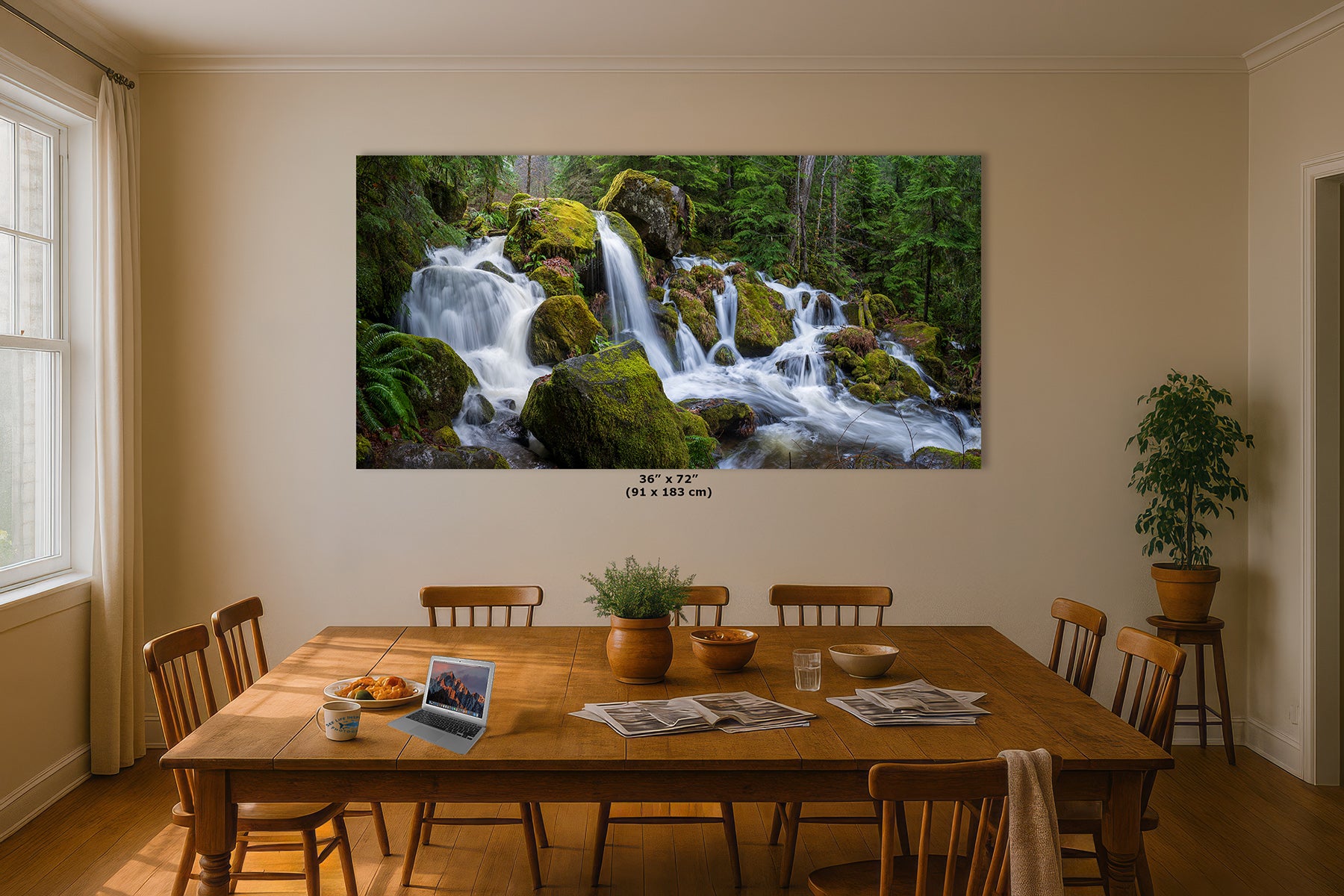 A large rectangular wall art print showing a forest waterfall cascading over mossy rocks, displayed above a wooden dining table in a real room with chairs, plants, and natural light from a window.