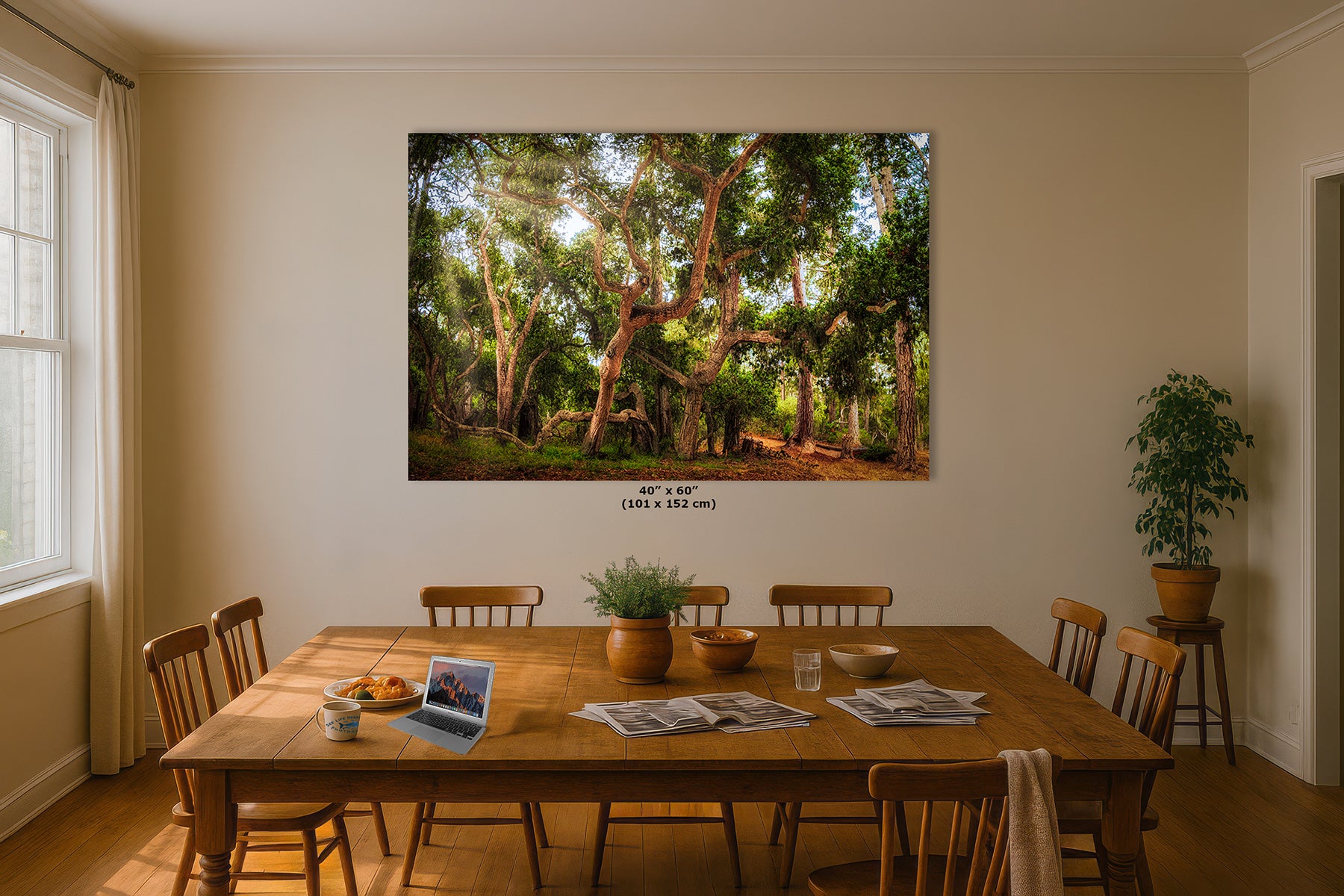 A large framed wall art print depicting a lush green coast live oak forest with sunlight filtering through trees, displayed on a beige wall above a wooden dining table in a styled room.