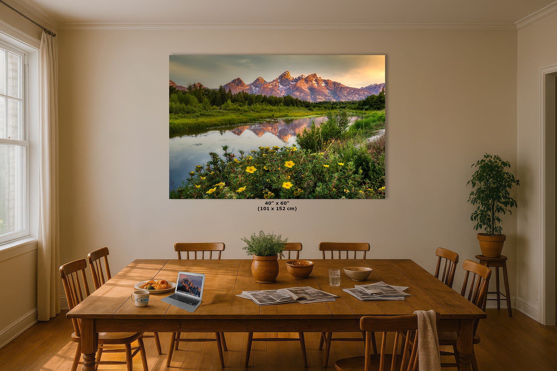 A large wall art print depicting a Grand Teton mountain landscape with a reflective lake and yellow wildflowers, displayed above a wooden dining table in a warm, modern room.