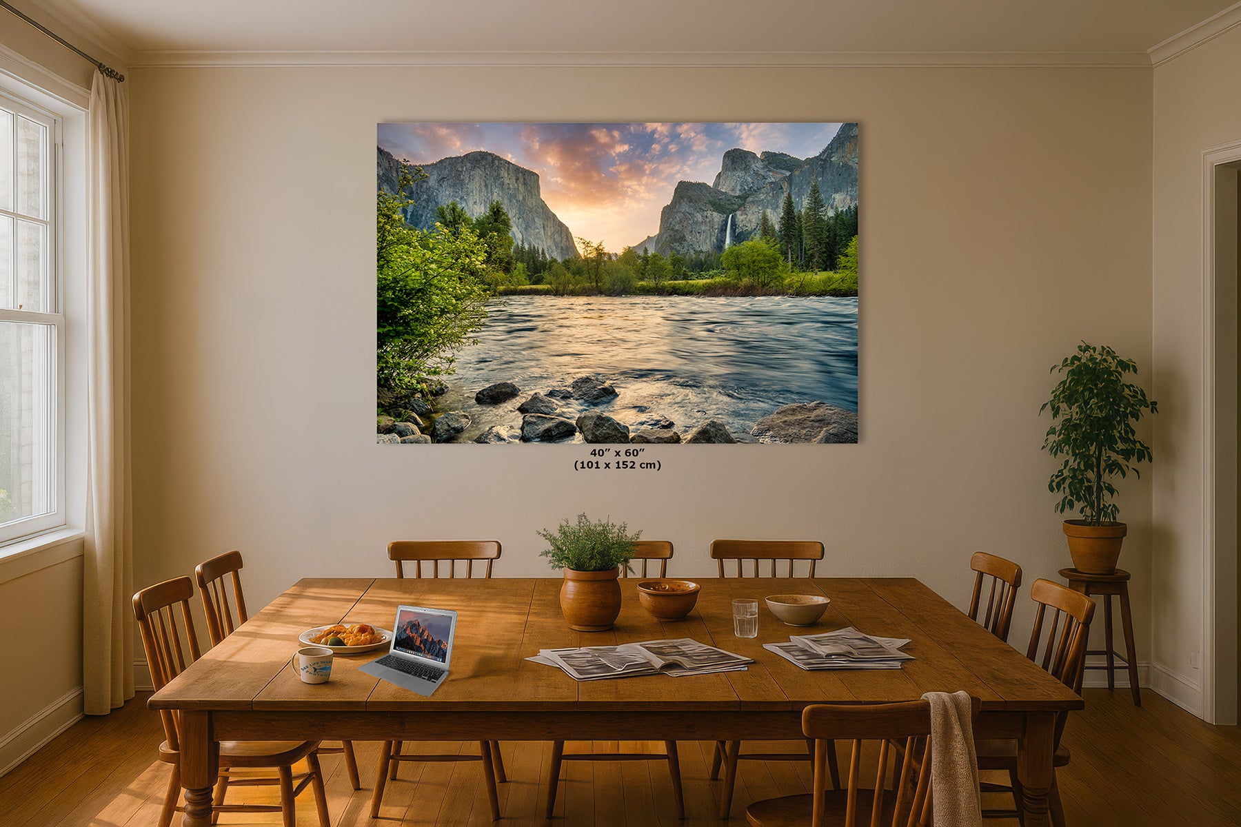 A large Yosemite wall art print showing a mountain river landscape with a sunset sky, displayed above a wooden dining table in a warm beige room with chairs, plants, and natural light from a window.
