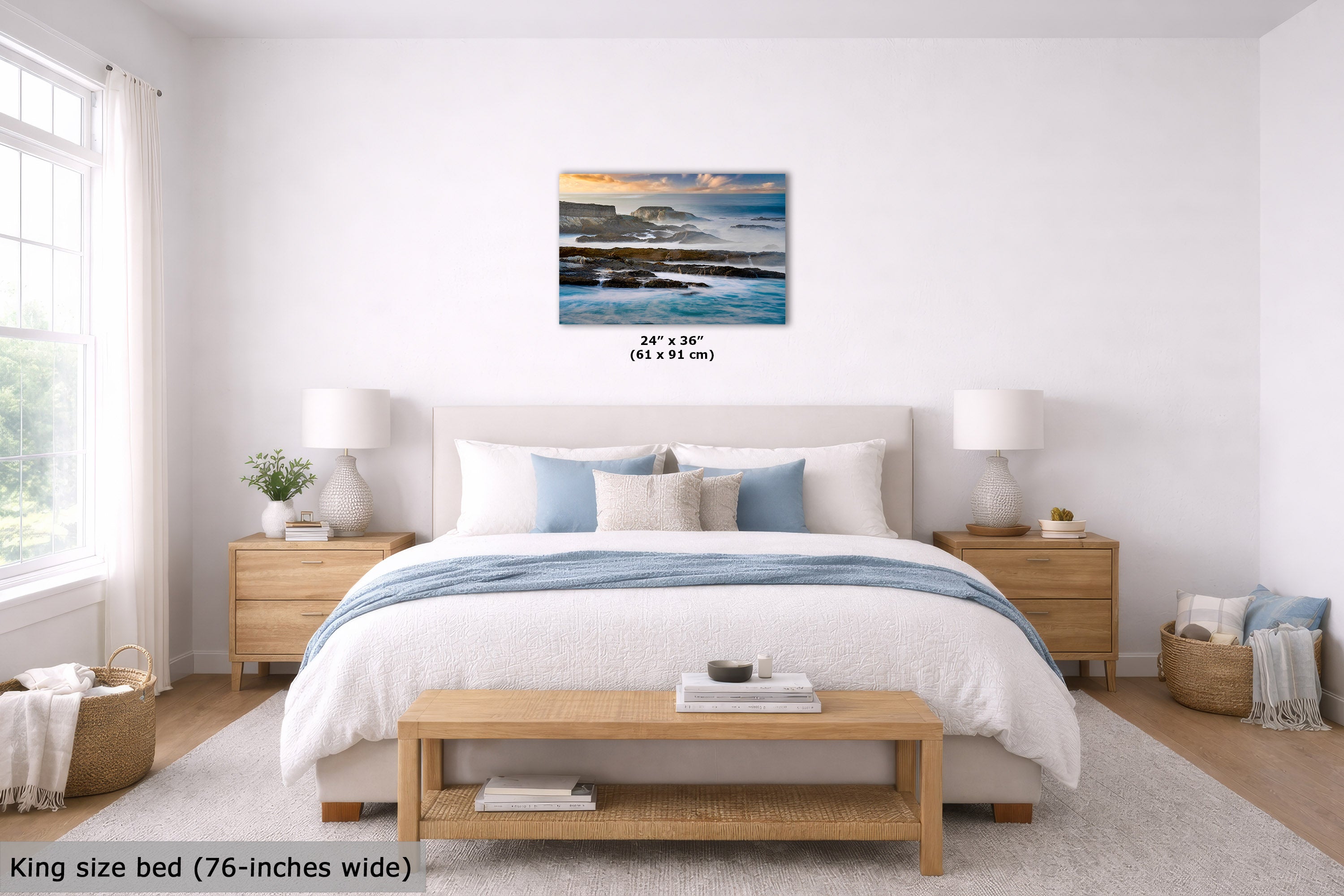 A coastal landscape wall art print hangs above a neatly made bed in a bright, modern bedroom. The print shows blue ocean water, dark rocky shoreline, and a warm sunset sky.