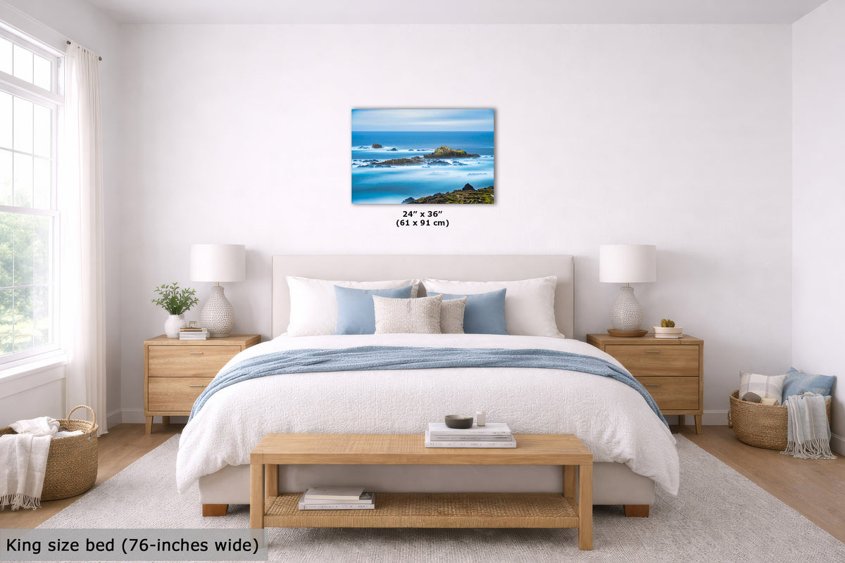A framed ocean landscape print hangs above a bed in a bright, modern bedroom. The artwork shows blue ocean, white foam, and dark rocks.