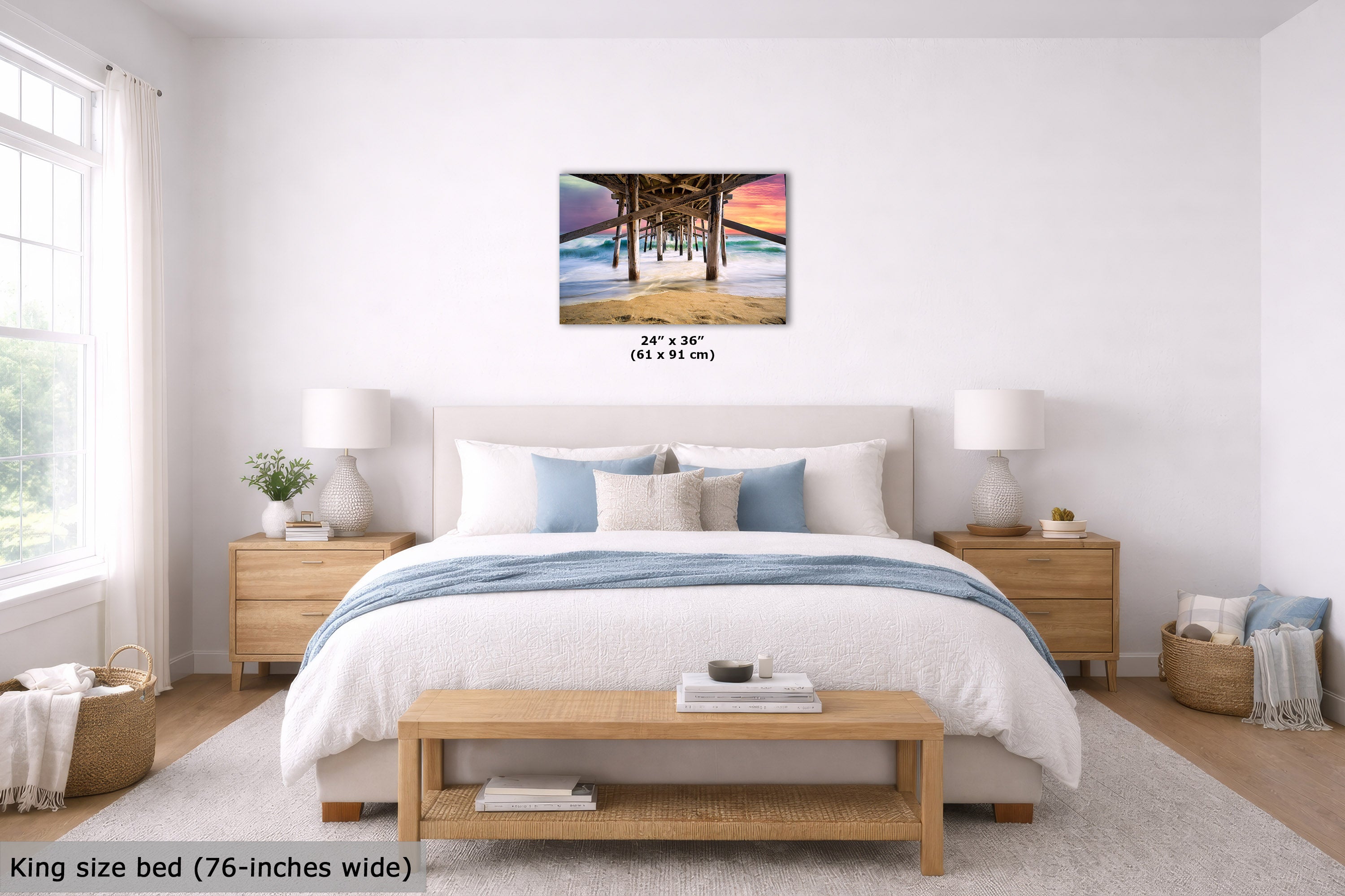 A wall art print showing a pier overlooking a beach at sunset, displayed above a neatly made bed in a bright, modern bedroom. The print includes size text reading 24" x 36" (61 x 91 cm).