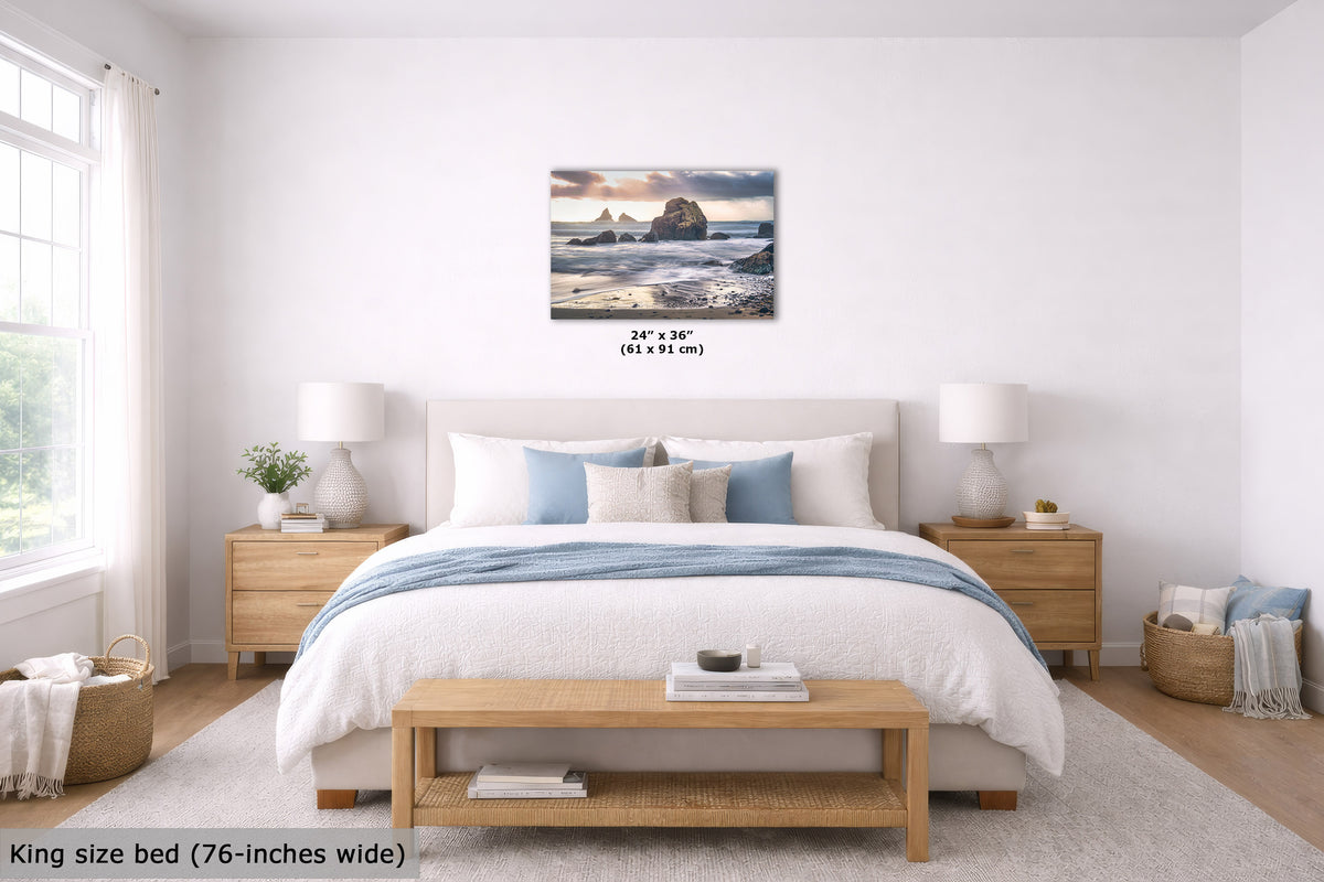 A wall art print depicting a rocky beach seascape with waves, shown hanging above a neatly made bed in a bright, modern bedroom. The print includes size text reading 24" x 36" (61 x 91 cm), and the room scene also includes a bed, nightstands, lamps, a bench, and a window.