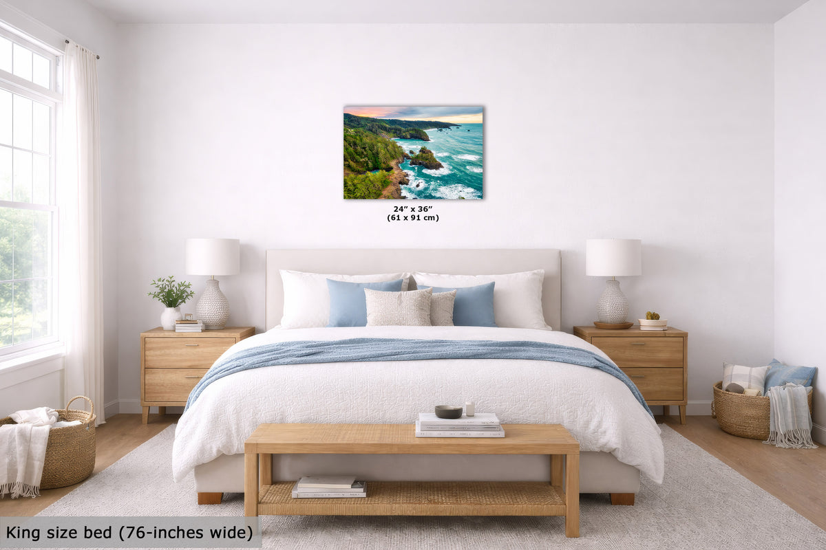 A coastal landscape wall art print hangs above a neatly made bed in a bright, modern bedroom. The artwork shows turquoise ocean waves crashing on rocky green cliffs under a pink and blue sunset sky.