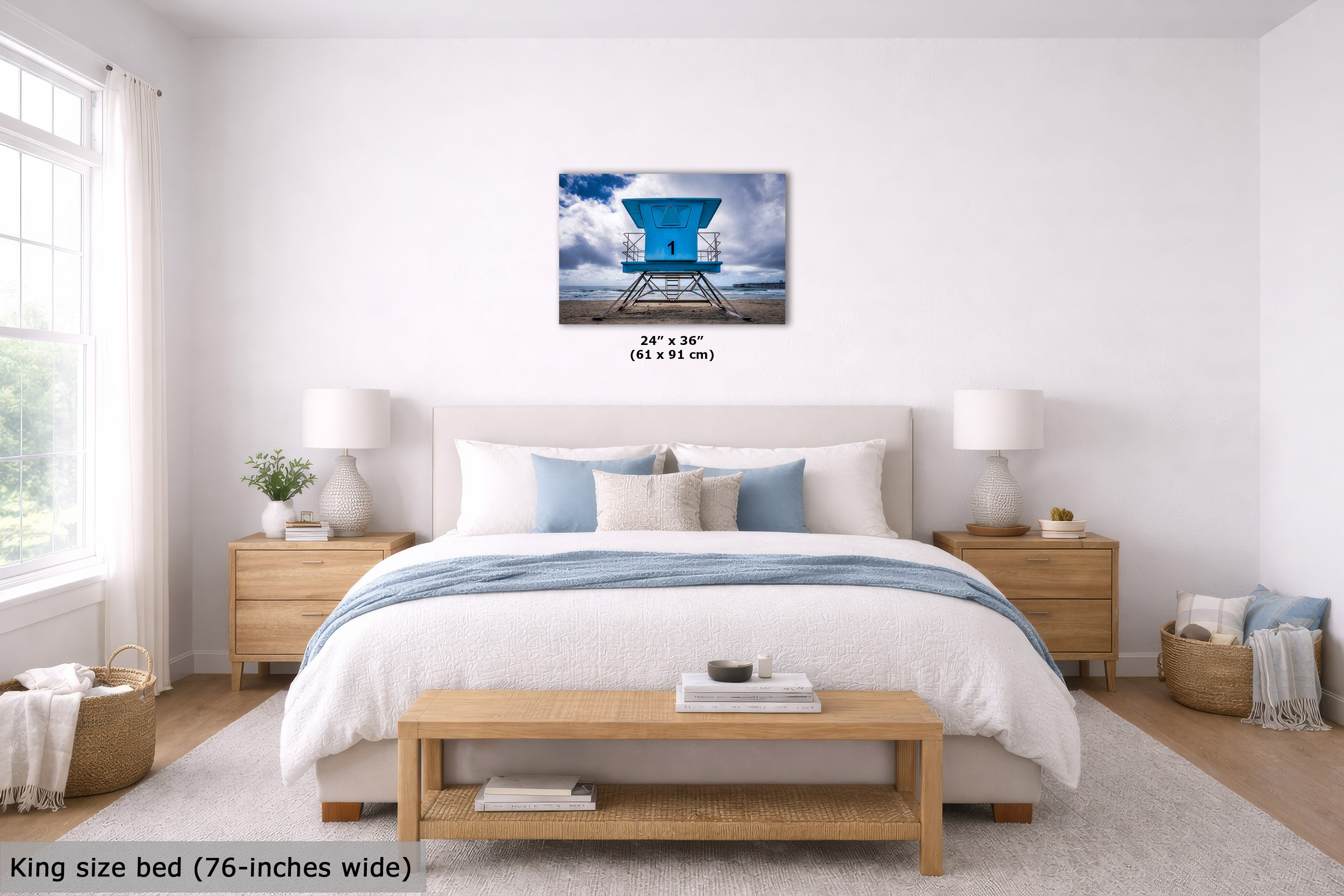 A wall art print showing a bright blue lifeguard tower over water, displayed above a neatly made bed in a bright, modern bedroom. The print includes size text reading 24" x 36" (61 x 91 cm).
