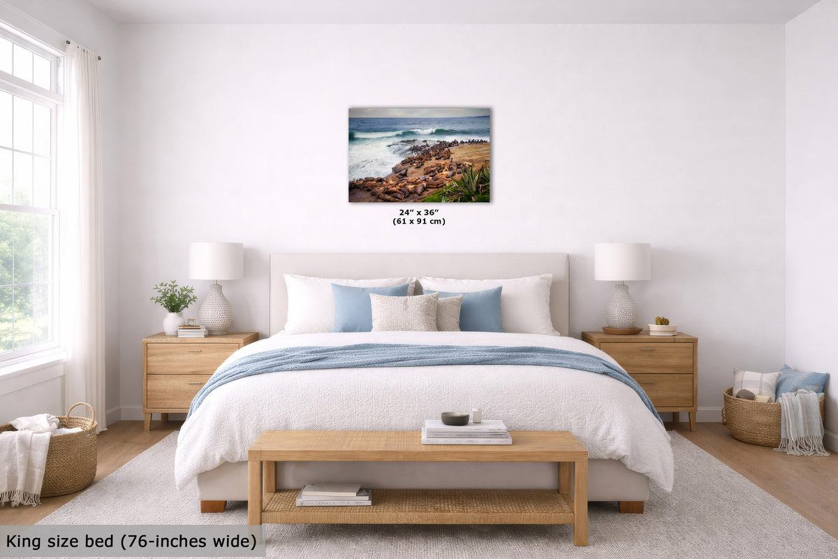 A coastal landscape wall art print hangs above a neatly made bed in a bright, modern bedroom. The print shows ocean waves crashing onto a rocky shore with warm sandy beach tones.