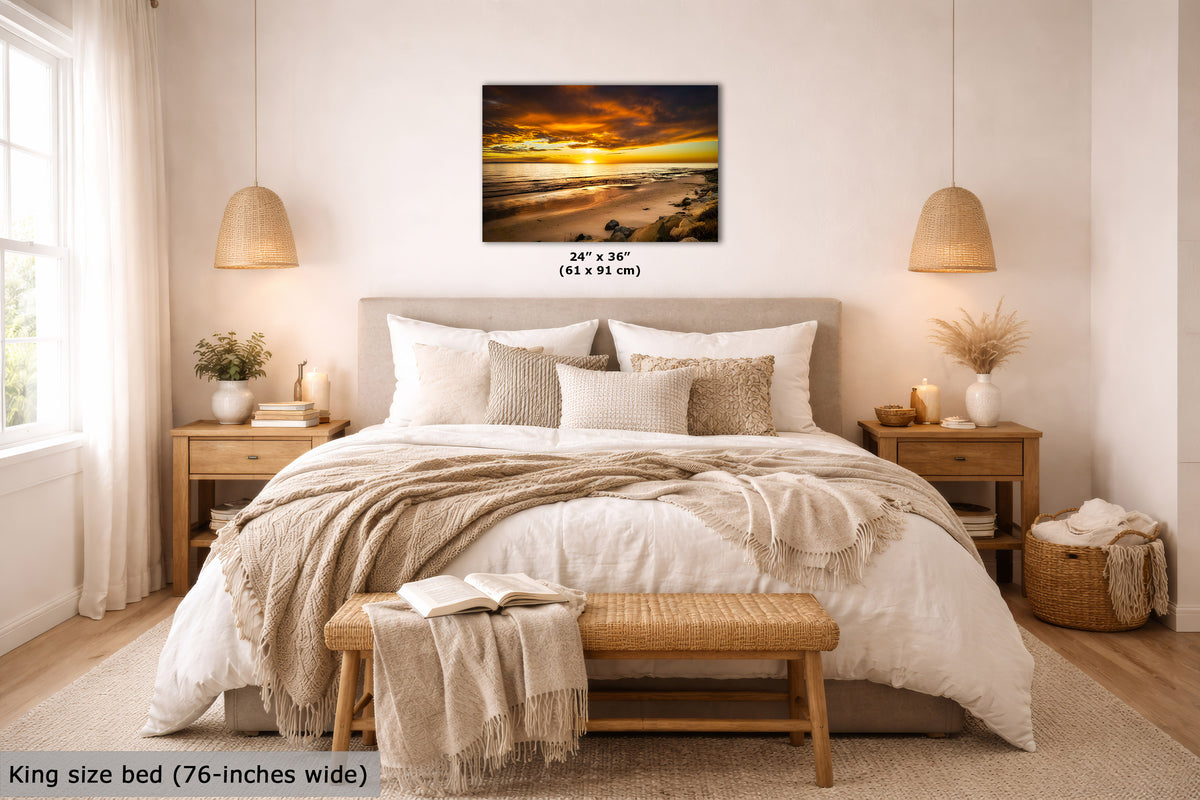 A wall art print showing a dramatic beach sunset in warm orange, yellow, and brown tones, displayed above a neatly made bed in a bright, modern bedroom. The print includes size text reading 24 x 36 inches (61 x 91 cm).