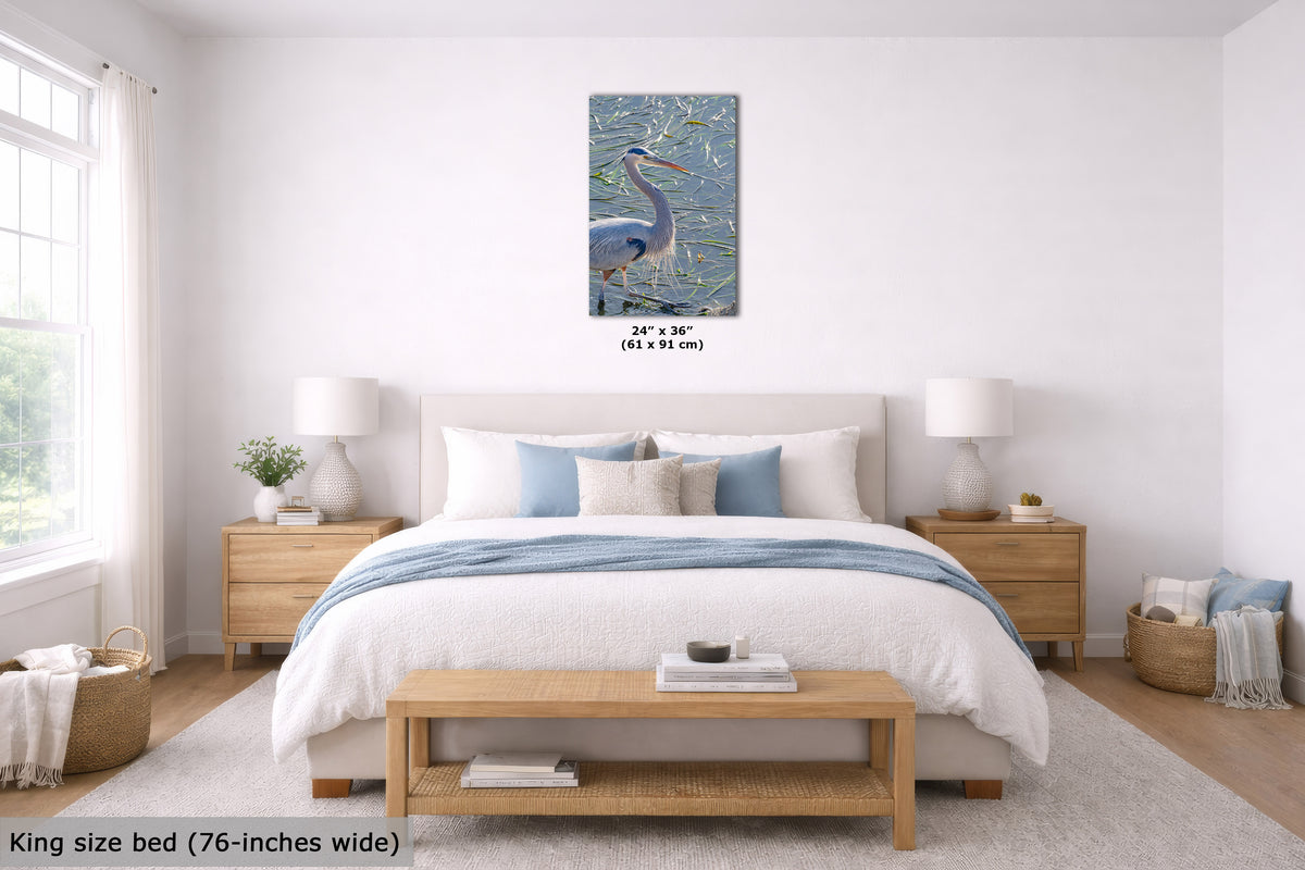 A bedroom scene showing a framed wall art print above a bed. The artwork depicts a blue heron in a marshy landscape with reeds and water.