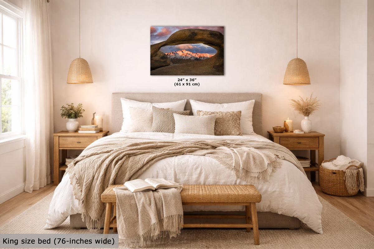 A wall art print showing a desert arch landscape at sunset is displayed above a neatly made bed in a bright, modern bedroom. The print includes size text reading 24 x 36" (61 x 91 cm), and the room scene includes a bed, nightstands, lamps, and decorative textiles.