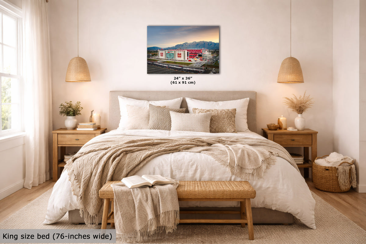 A wall art print showing a colorful mountain landscape with the 49er's Levi's Stadium, displayed above a neatly made bed in a bright, modern bedroom. The print includes size text reading 24 x 36 inches (61 x 91 cm), and the room scene also includes a bed, pillows, lamps, nightstands, and a bench.
