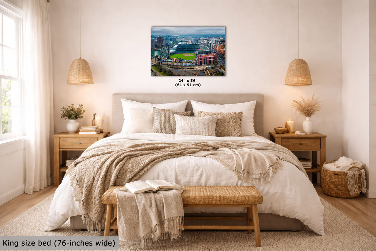 A San Francisco cityscape wall art print showing the Giants' stadium and surrounding skyline, displayed above a neatly made bed in a bright, modern bedroom. The print includes size text reading 24" x 36" (61 x 91 cm), and the scene is shown in a styled interior with bedding, lamps, nightstands, and a bench.