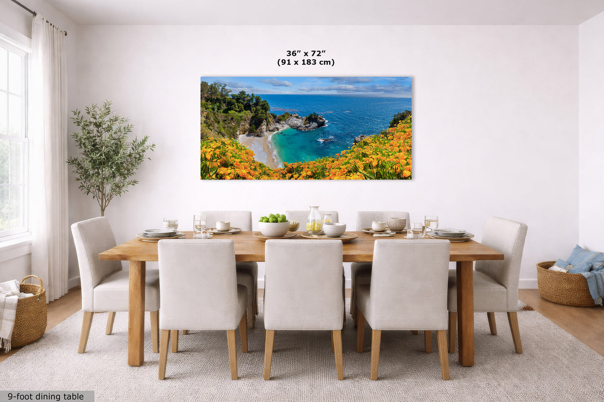 A large rectangular coastal landscape wall art print hangs above a dining table in a bright modern dining room. The artwork shows a sunny beach cove with turquoise water, rocky cliffs, green trees, and bright orange flowers.