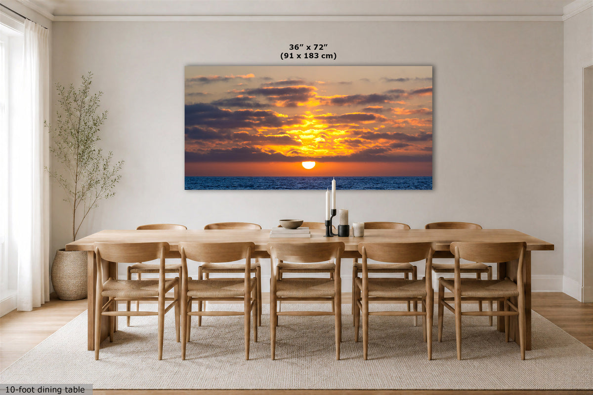 A large rectangular wall art print showing a dramatic sunset over the ocean, displayed above a wooden dining table and chairs in a bright modern dining room. The print includes size text at the top reading 36 x 72 (91 x 183 cm).