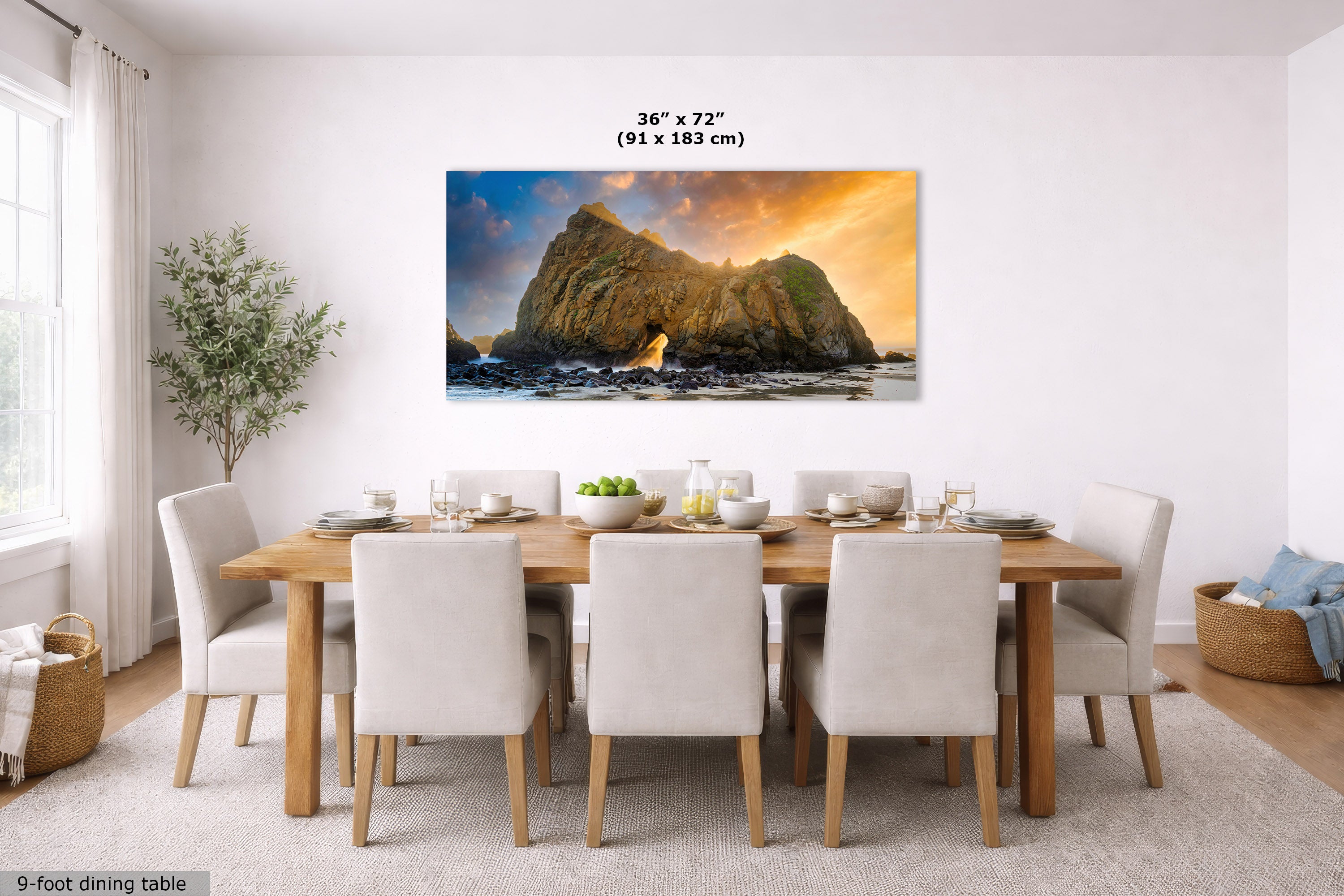 A large rectangular wall art print showing a dramatic rocky coastline at sunset, displayed above a wooden dining table with white upholstered chairs in a bright dining room. The print includes size text reading 36 x 72 inches (91 x 183 cm).