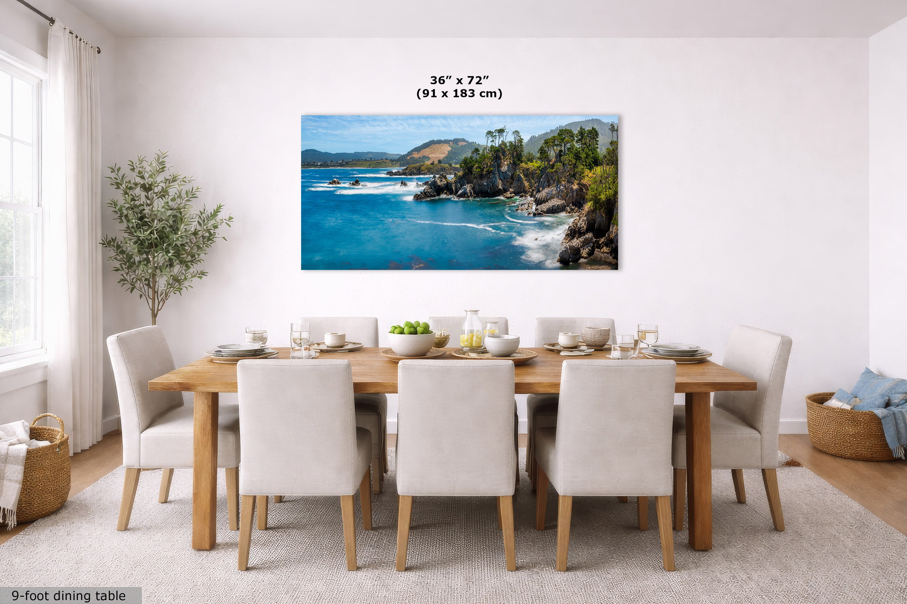 A coastal landscape wall art print hangs above a wooden dining table with upholstered chairs in a bright dining room. The print depicts a rocky blue-water shoreline with green cliffs and sky.