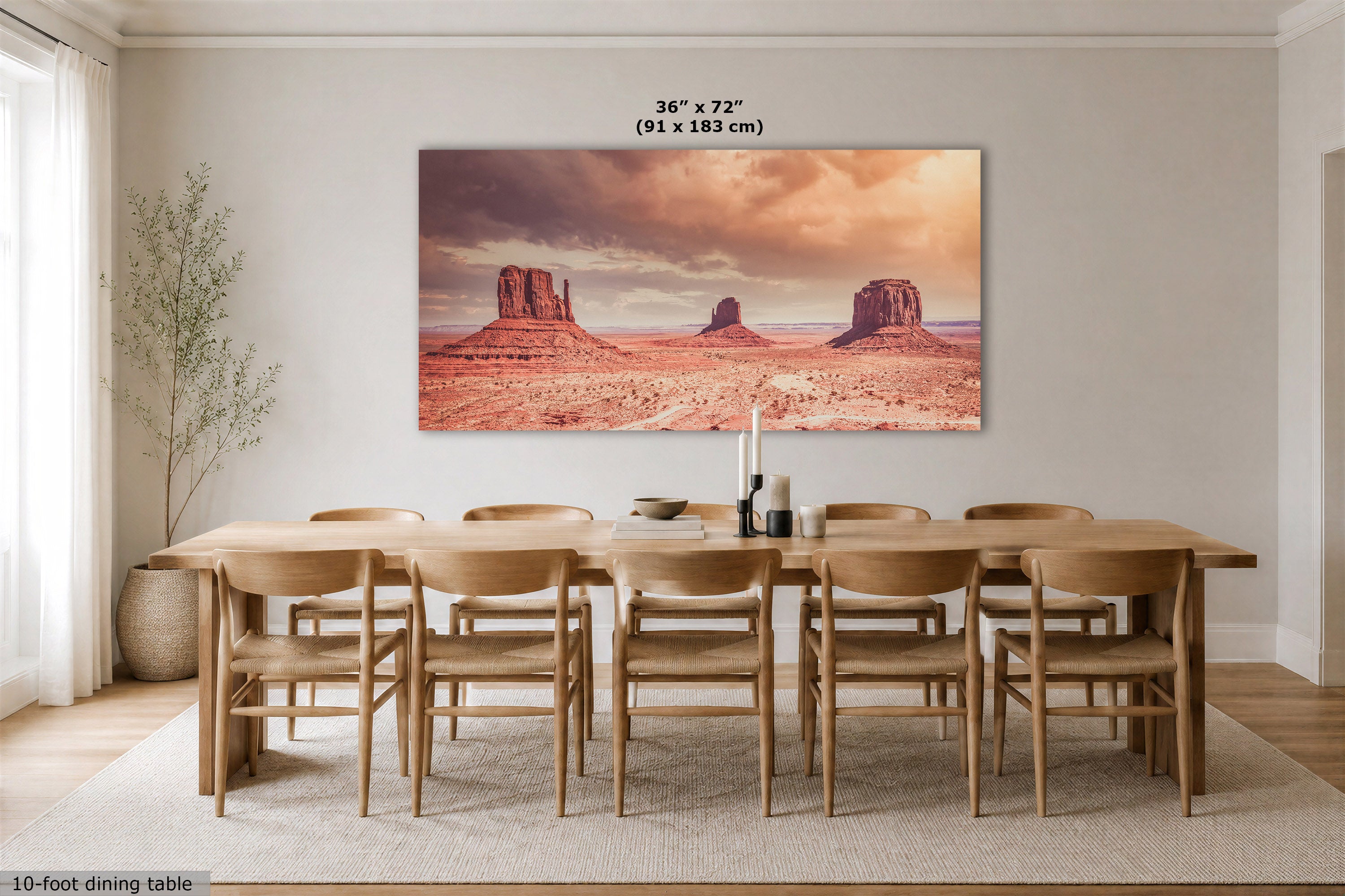 A large rectangular wall art print showing a desert landscape with rock buttes under a dramatic sky, displayed above a wooden dining table and chairs in a bright dining room. Text above the print indicates dimensions 36 x 72 in (91 x 183 cm).