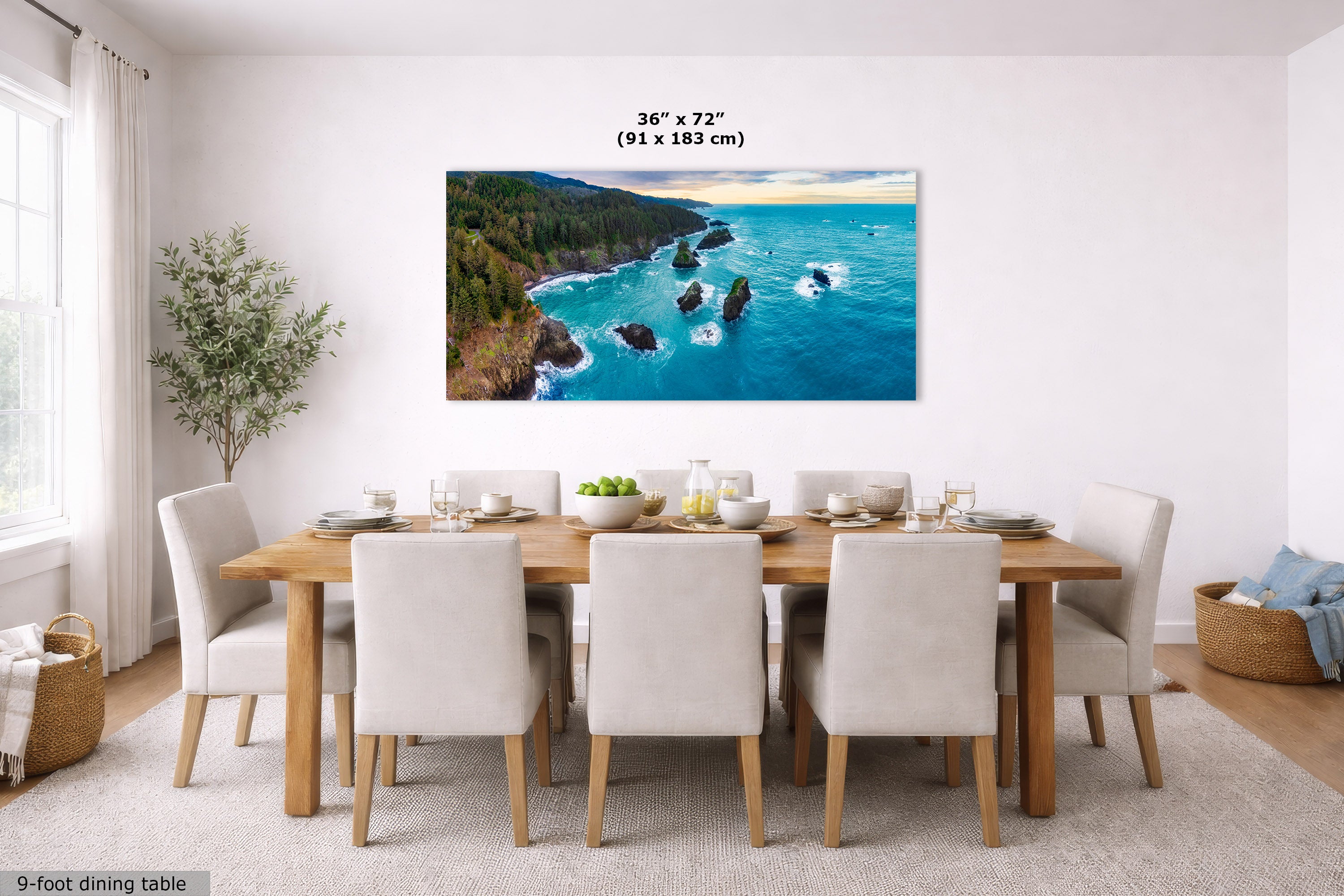 A large rectangular coastal landscape wall art print hangs above a dining table in a bright modern dining room. The artwork shows turquoise water, rocky shoreline, green forest, and a pale sky.
