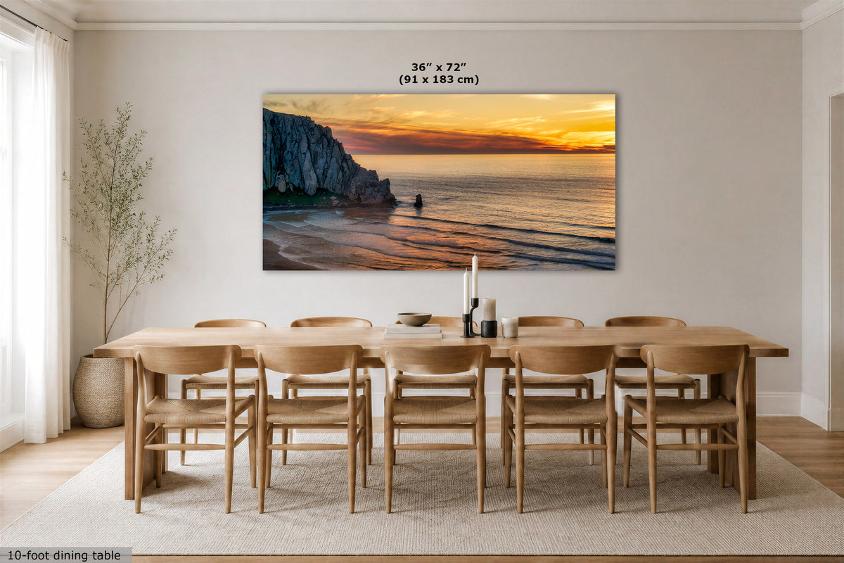 A large rectangular wall art print showing a coastal cliff and beach sunset hangs above a wooden dining table and chairs in a bright modern dining room. The print includes size text reading 36 x 72 (91 x 183 cm).