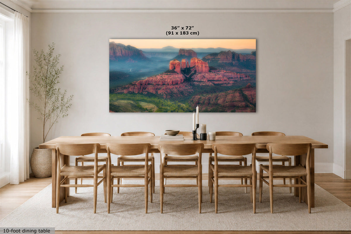 A large rectangular wall art print showing a desert canyon landscape with red rock formations and blue mist hangs above a wooden dining table and chairs in a bright neutral dining room. Small text above the artwork reads 36 x 72 (91 x 183 cm).
