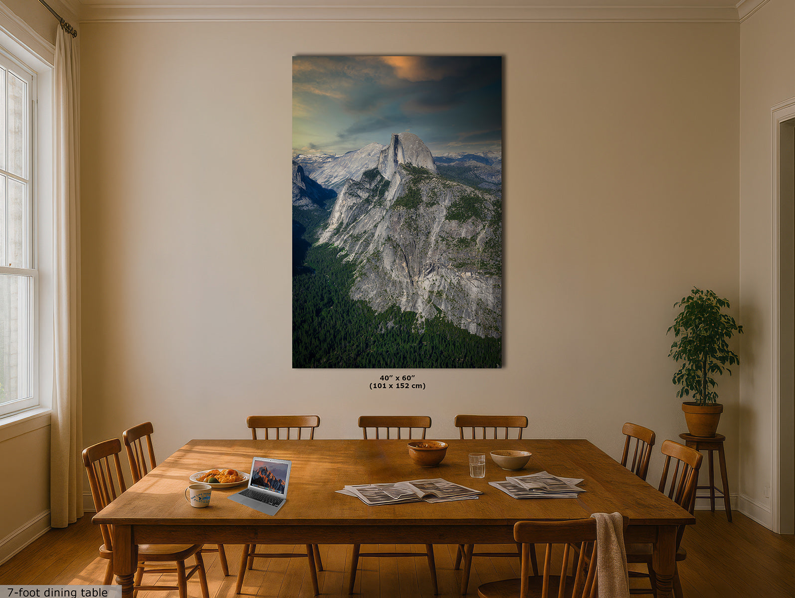 A large vertical wall art print of Half Dome in Yosemite, depicting a mountain landscape with a glowing sunset sky, displayed above a wooden dining table in a beige dining room with chairs and a potted plant.