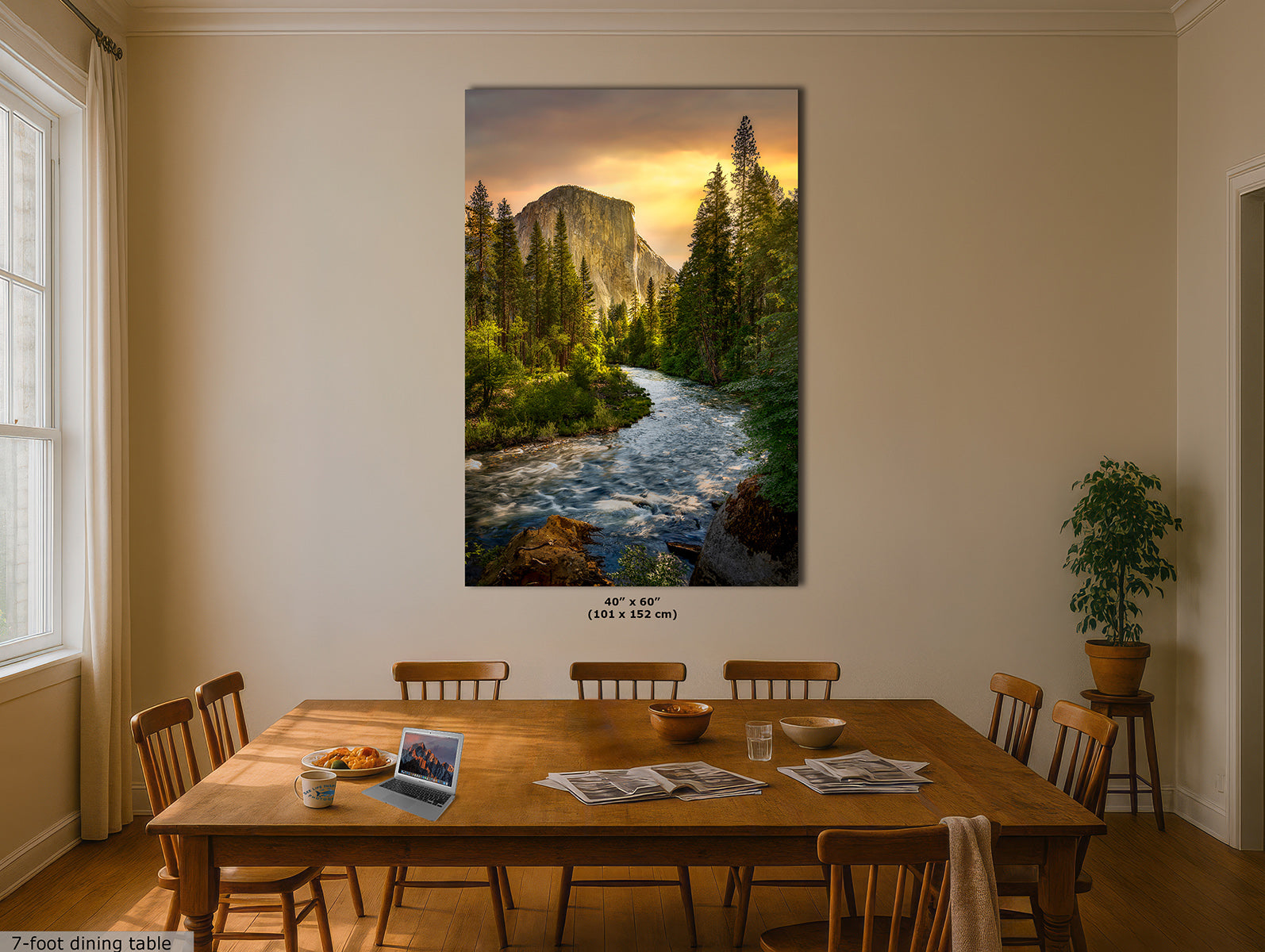 A large vertical Yosemite wall art print of El Capitain and the Merced River that hangs on a beige wall above a wooden dining table in a dining room. The artwork shows a rushing river, dense evergreen trees, and a glowing sunset sky. Small text at the bottom reads 40 x 60-inches, and a small label at bottom left says 7-foot dining table.